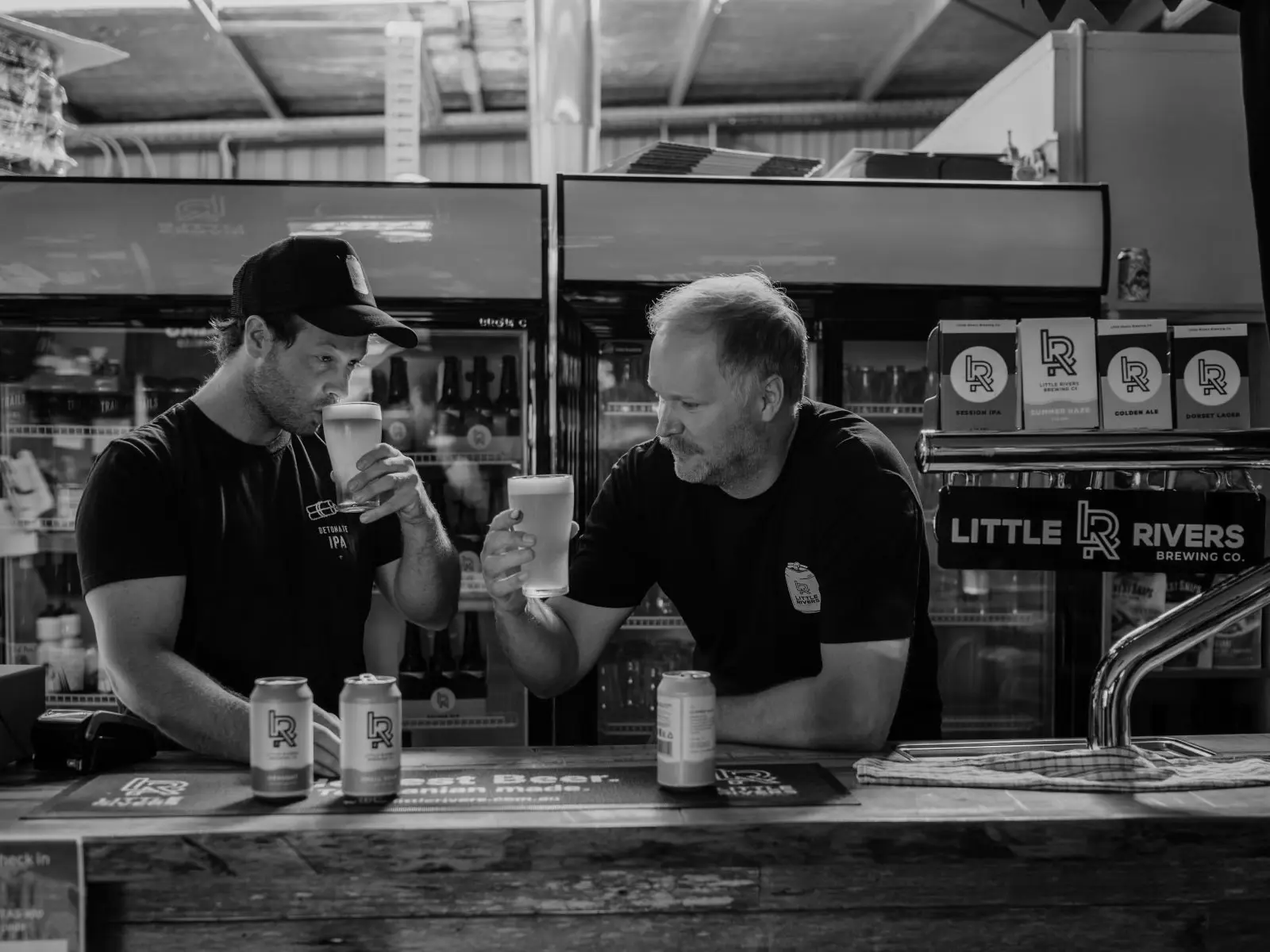 Two men stand behind a wooden bar of a brewery, drinking glasses of beer. Cans and bottles are on display and the taps read 'Little Rivers'.