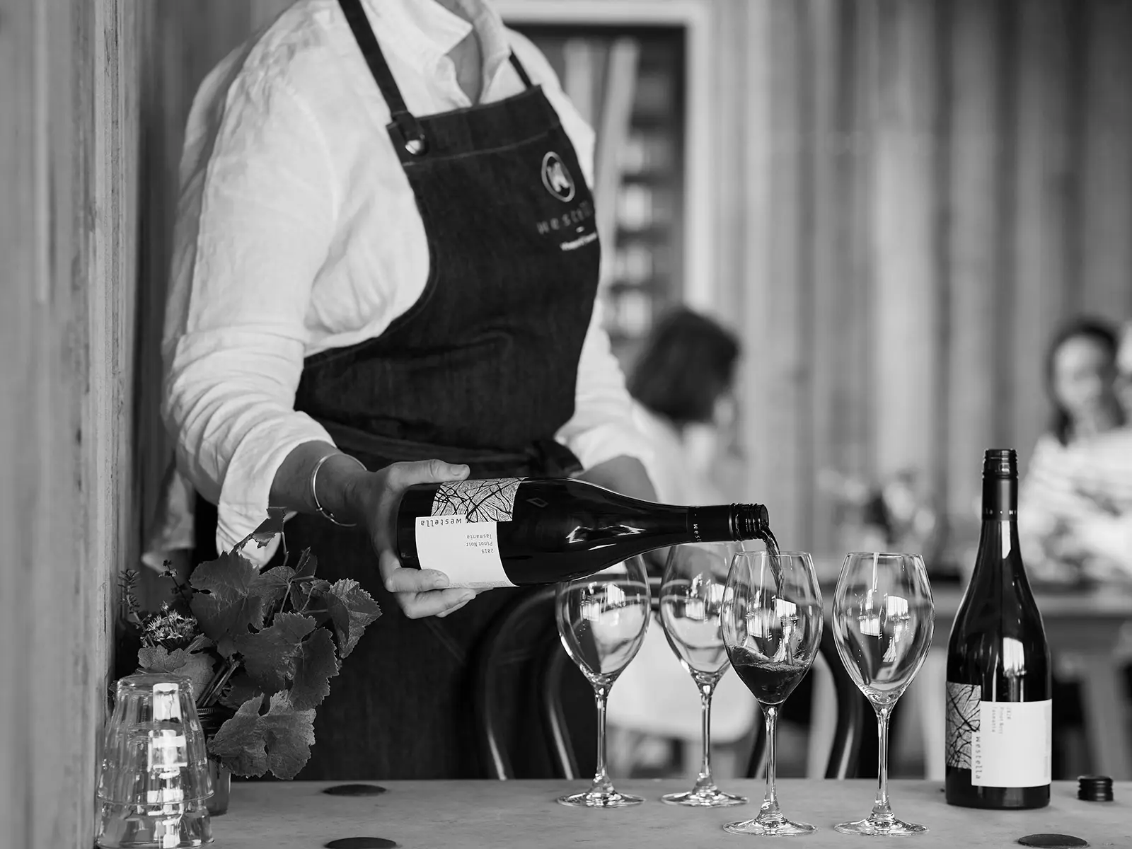Black and white image of a server in white shirt and apron, pouring red wine into lined up glasses.