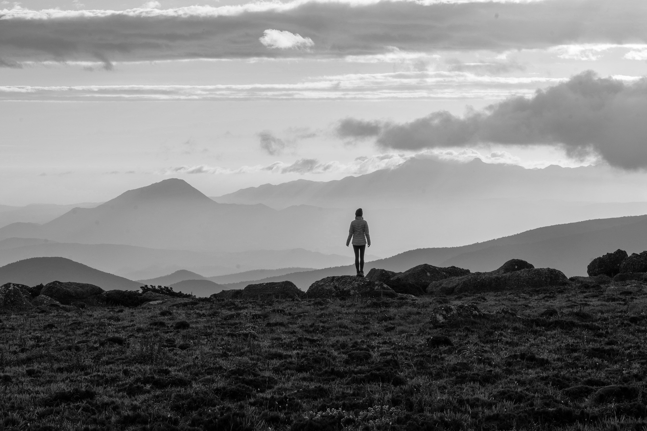 A solitary figure stands on rocky ground, gazing out across layers of misty mountain ranges under a moody, cloud-streaked winter sky.