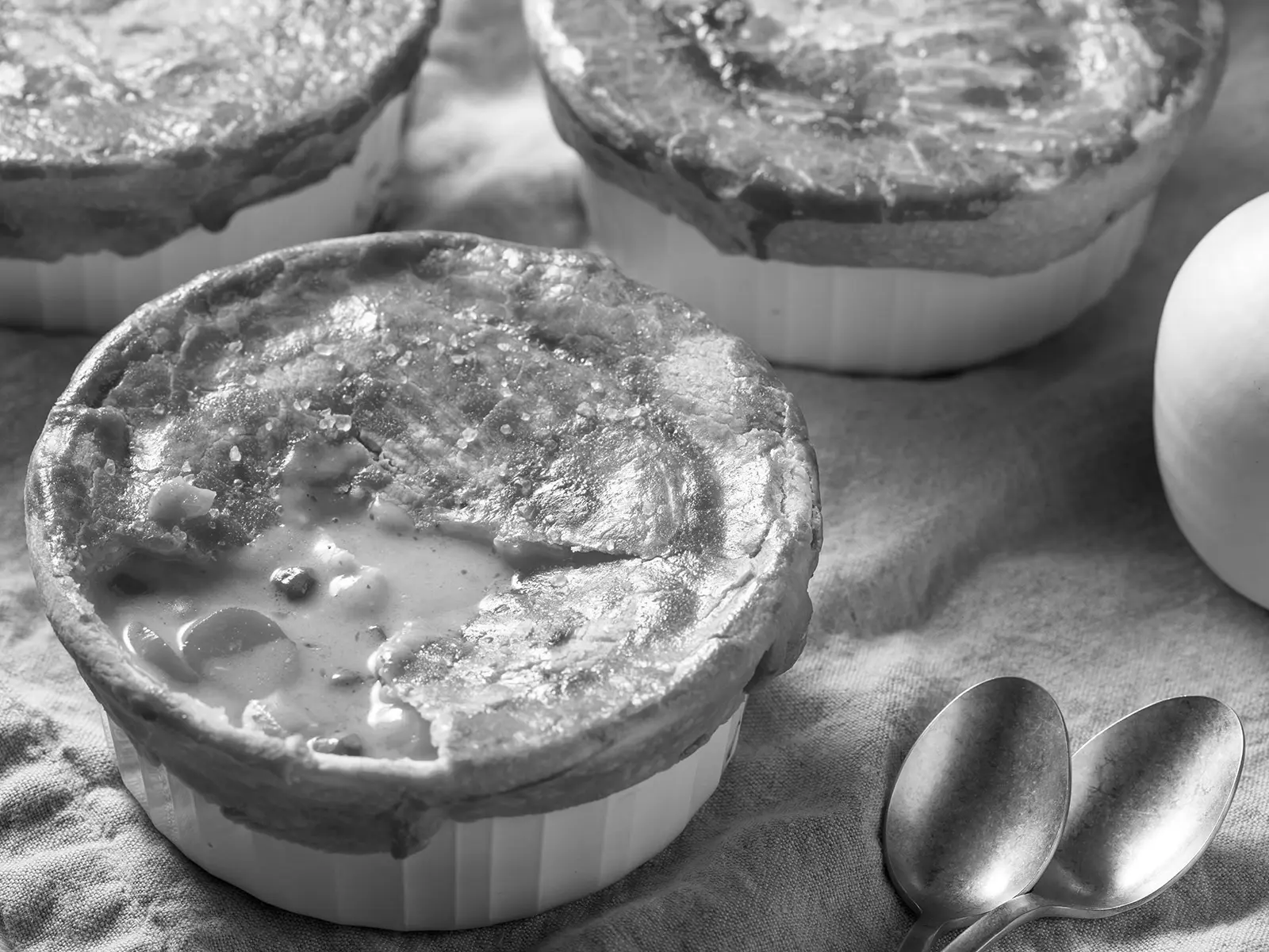 A close-up of individual pot pies in ramekins, topped with crispy pastry. One is cracked and the creamy filling is visible.