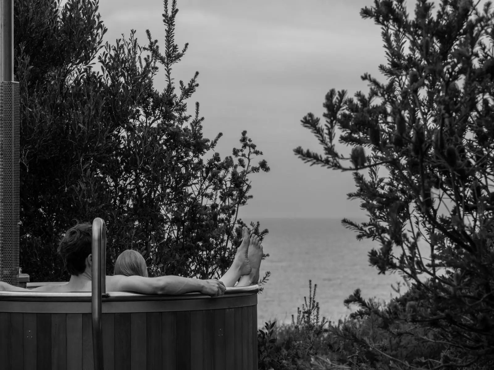 Two people relax in a large outdoor tub, their feet up on the edges. The view to the ocean beyond is framed by trees.