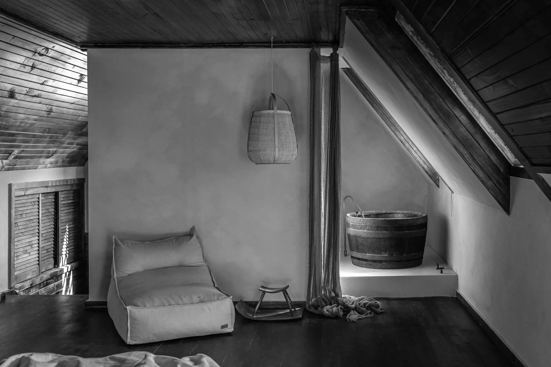Black and white image of a minimalistic indoor space with a wooden barrel bathtub, a cushioned lounge chair, a rocking footstool, a pendant light hanging from the ceiling, wooden paneling on the ceiling, and a curtain partially drawn to the floor.