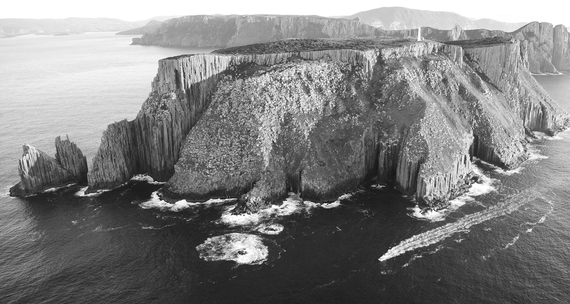An aerial black and white view over the Tasman Peninsula, with dramatic dolerite cliffs and waves crashing. A small boat powers through the water at the foot of the cliffs.