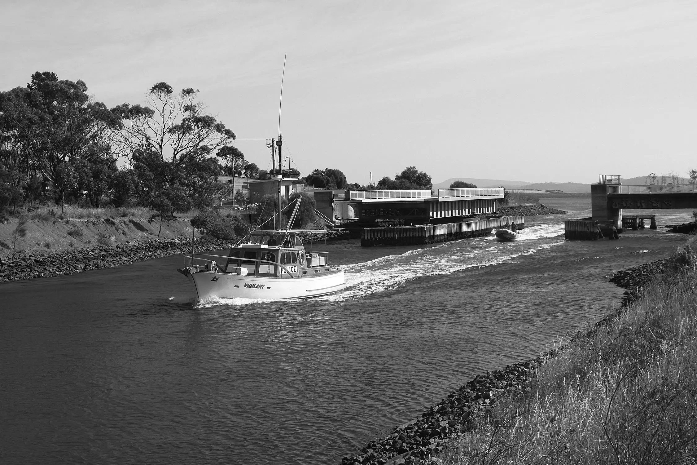 A small yacht cruises through an open bridge over a river, with bush lining the banks.