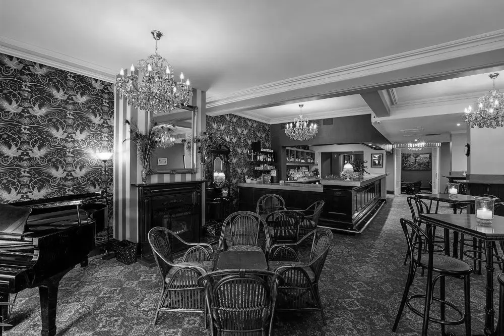 The interior of a historic bar, featuring chandeliers, ornate wallpaper and carpet, and a long bar. A piano stands to one side.