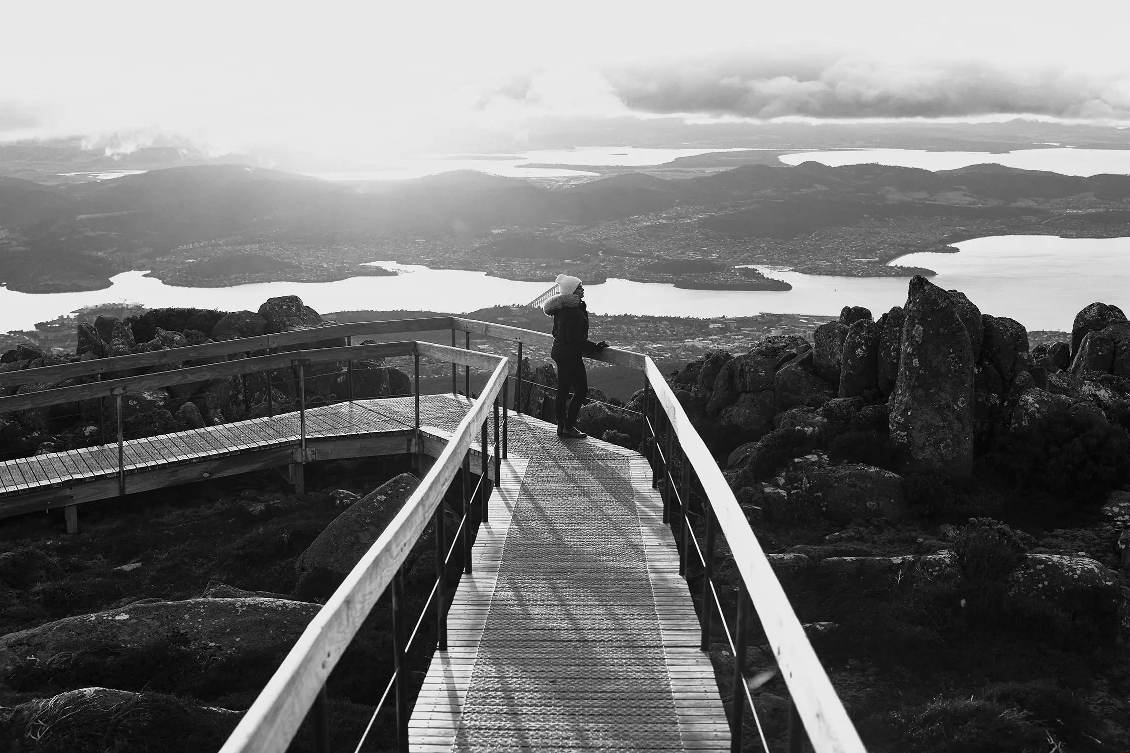 A woman stands at the end of a wooden lookout on top of a mountain, looking down over Hobart.