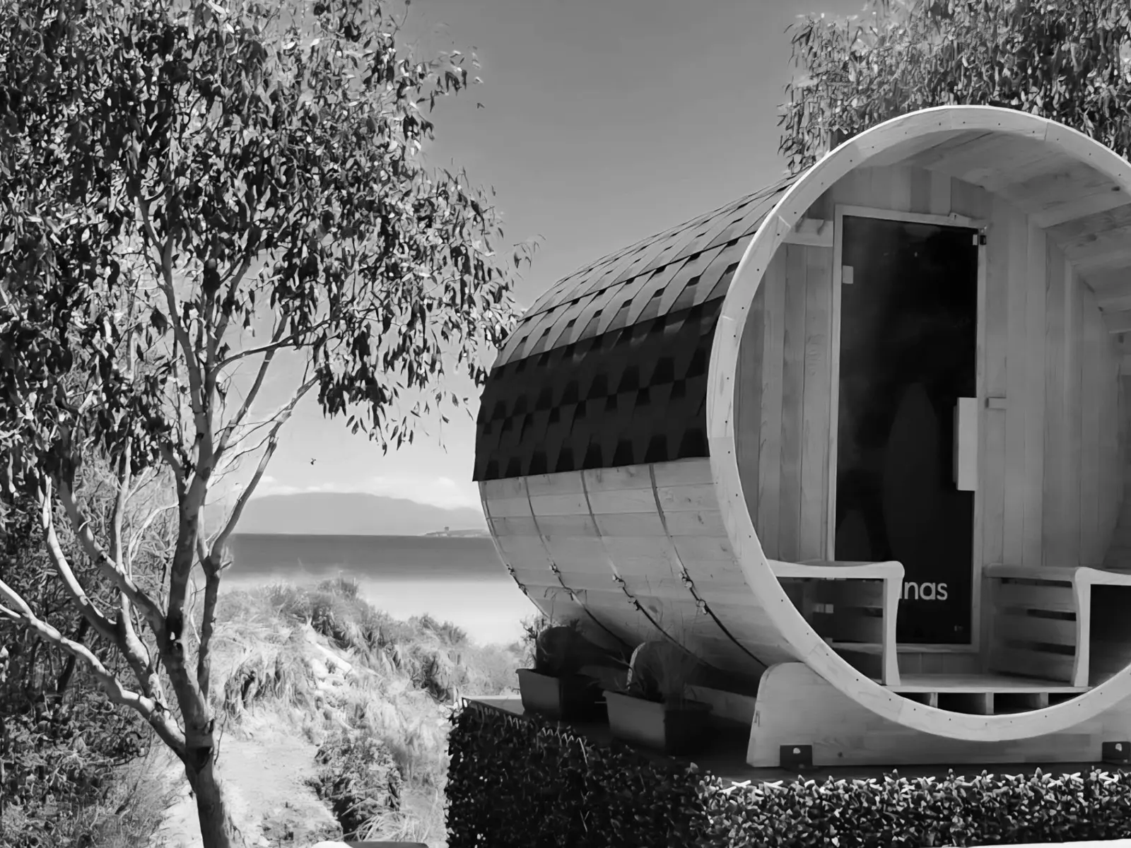 A barrel-shaped sauna sits atop a platform, looking out over a scenic beach-side bush view, framed by eucalyptus trees.