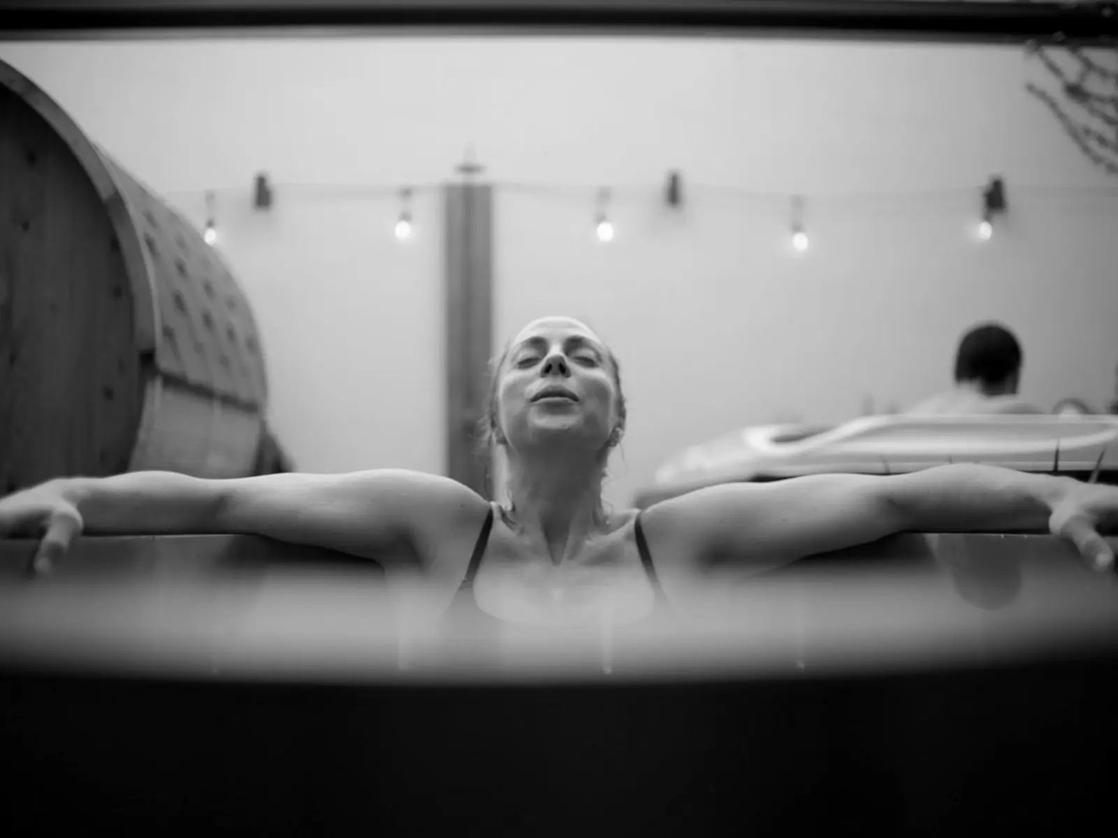Looking up over the edge of an outdoor tub, while a woman in bathers relaxes with her arms stretched out over the edges.