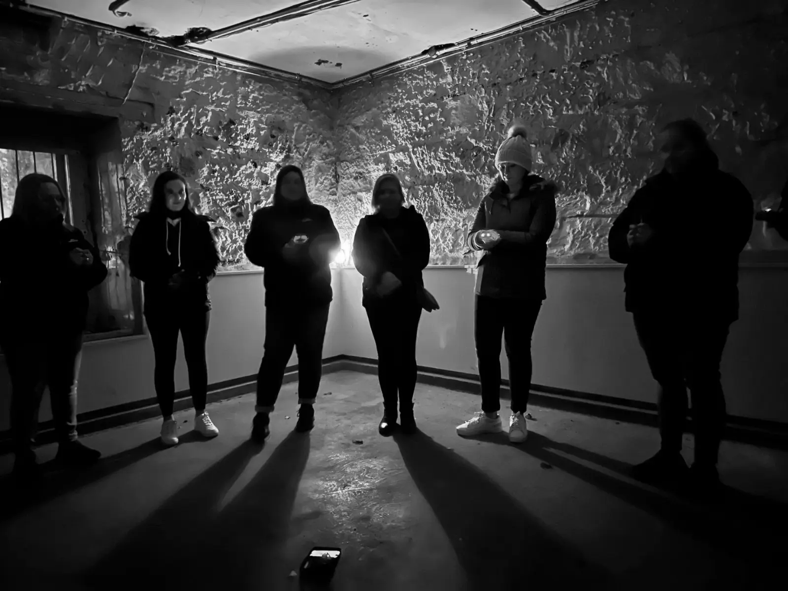 A group of people stand in an empty room of an old sandstone building, the lighting from behind throwing long, ominous shadows.