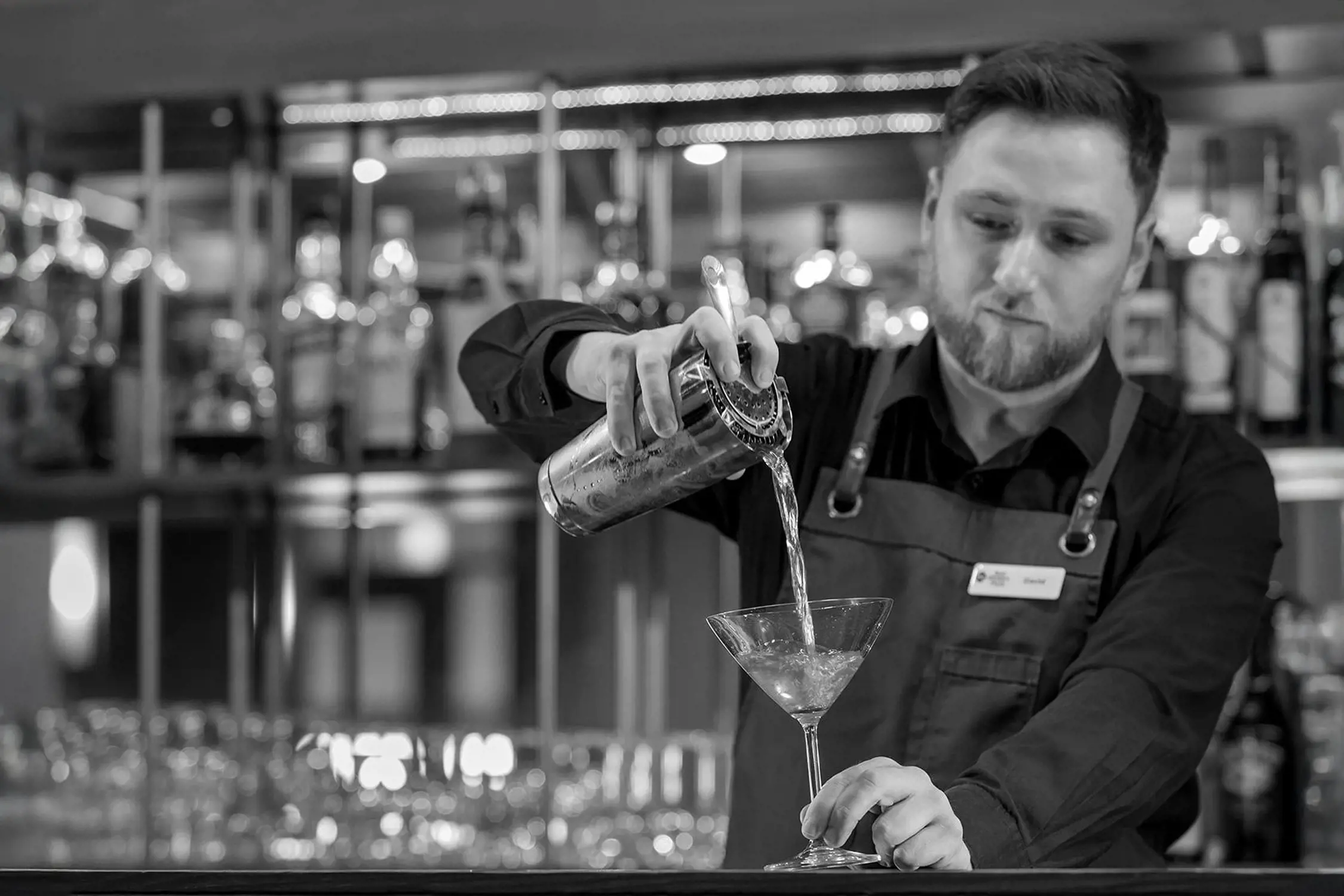 A focused bartender wearing a dark shirt and leather-strap apron pours a clear liquid from a metal cocktail shaker through a strainer into a martini glass on the bar counter. Behind him, shelves are lined with various bottles of liquor, softly illuminated by warm lighting.