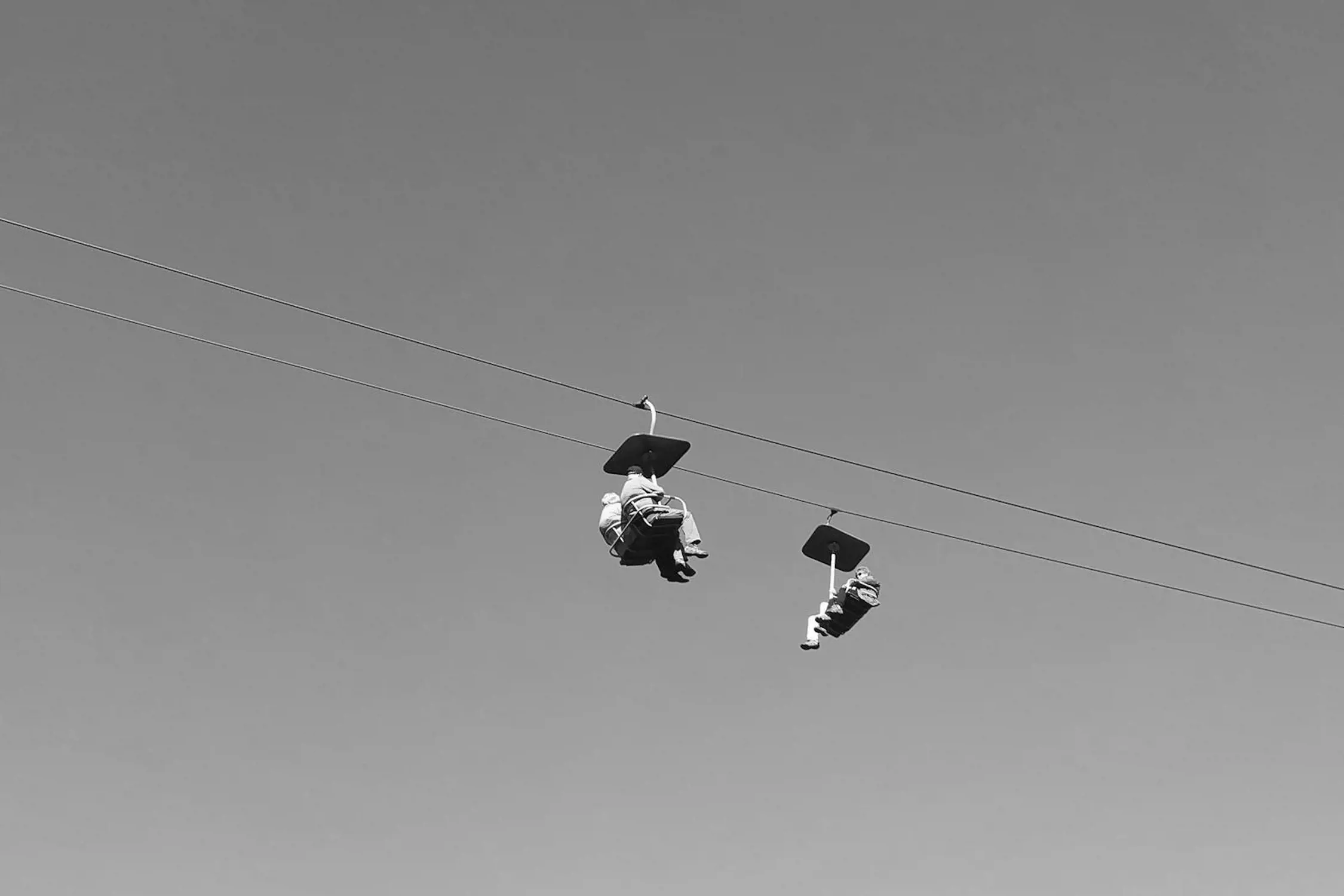Two people are seated on an open-air chairlift suspended high above the ground, slowly moving along thin cables against a clear sky. Each chair includes an overhead canopy for shade, and the individuals are positioned comfortably as they enjoy the elevated ride, hinting at a nearby recreational or ski area.