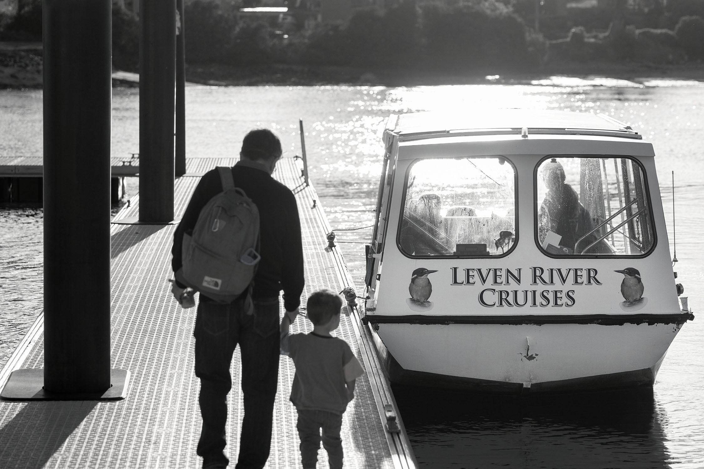 A man holds the hand of a small child as they walk along a jetty, towards a little river cruise boat emblazoned with the words 'Leven River Cruises'.