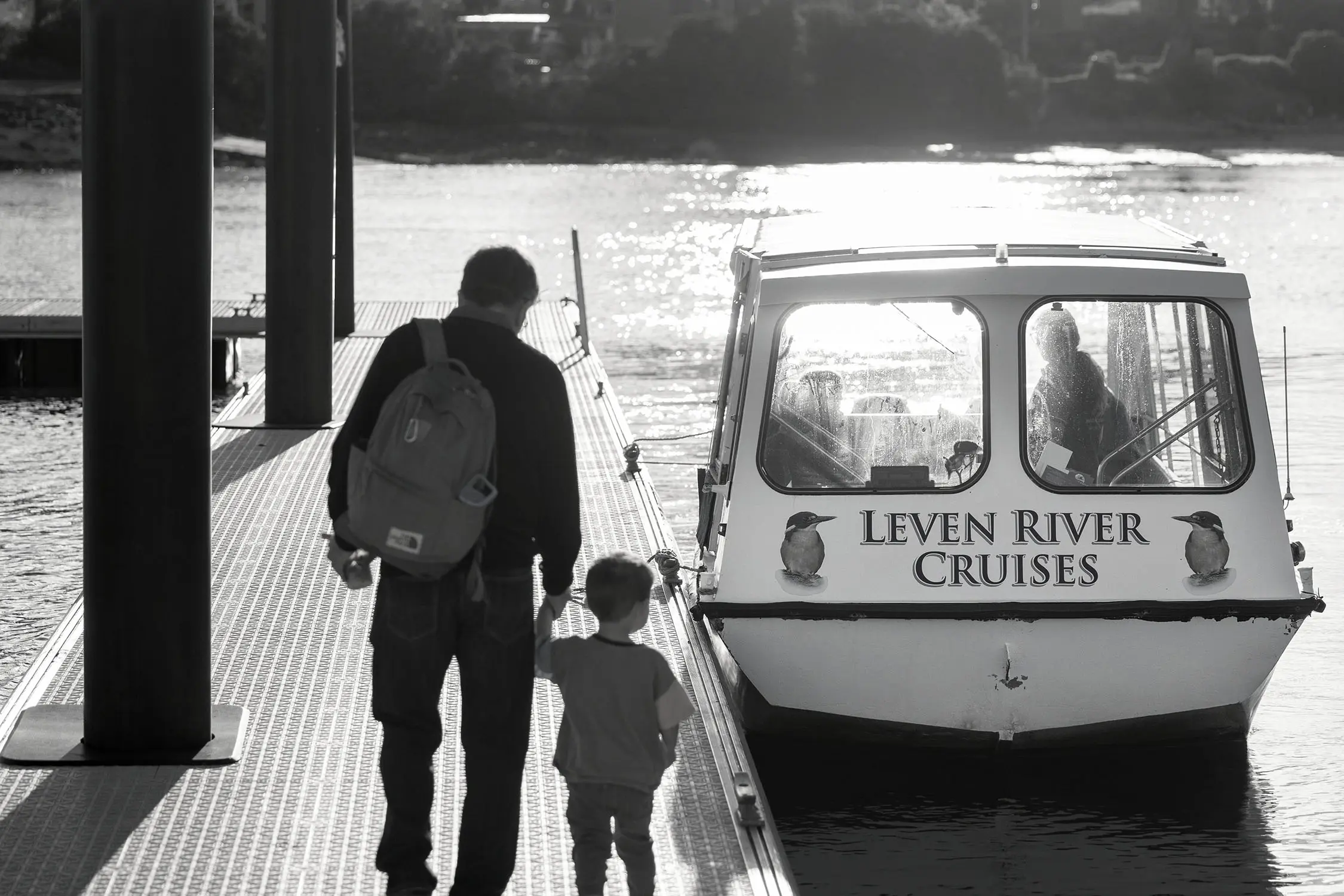 A man holds the hand of a small child as they walk along a jetty, towards a little river cruise boat emblazoned with the words 'Leven River Cruises'.