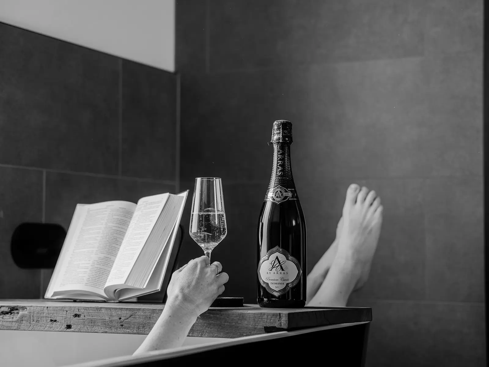 A person's feet stick out from the end of a bathtub. Across the tub, a bath caddy holds a bottle of wine, a wine glass and an open book.