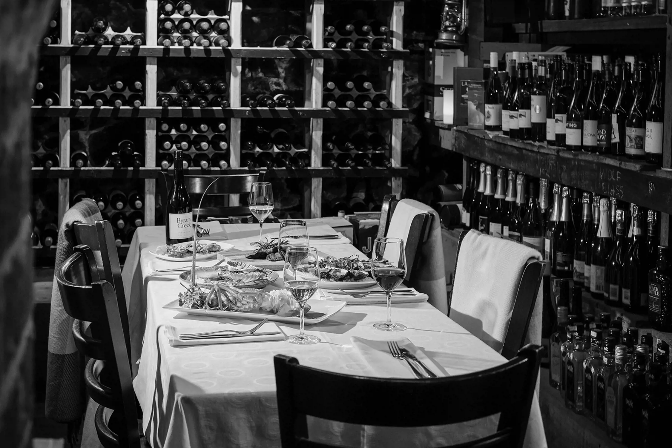 A cozy dining table set with plates of food, wine glasses, and bottles of wine on shelves behind the table in a rustic, dimly lit room.