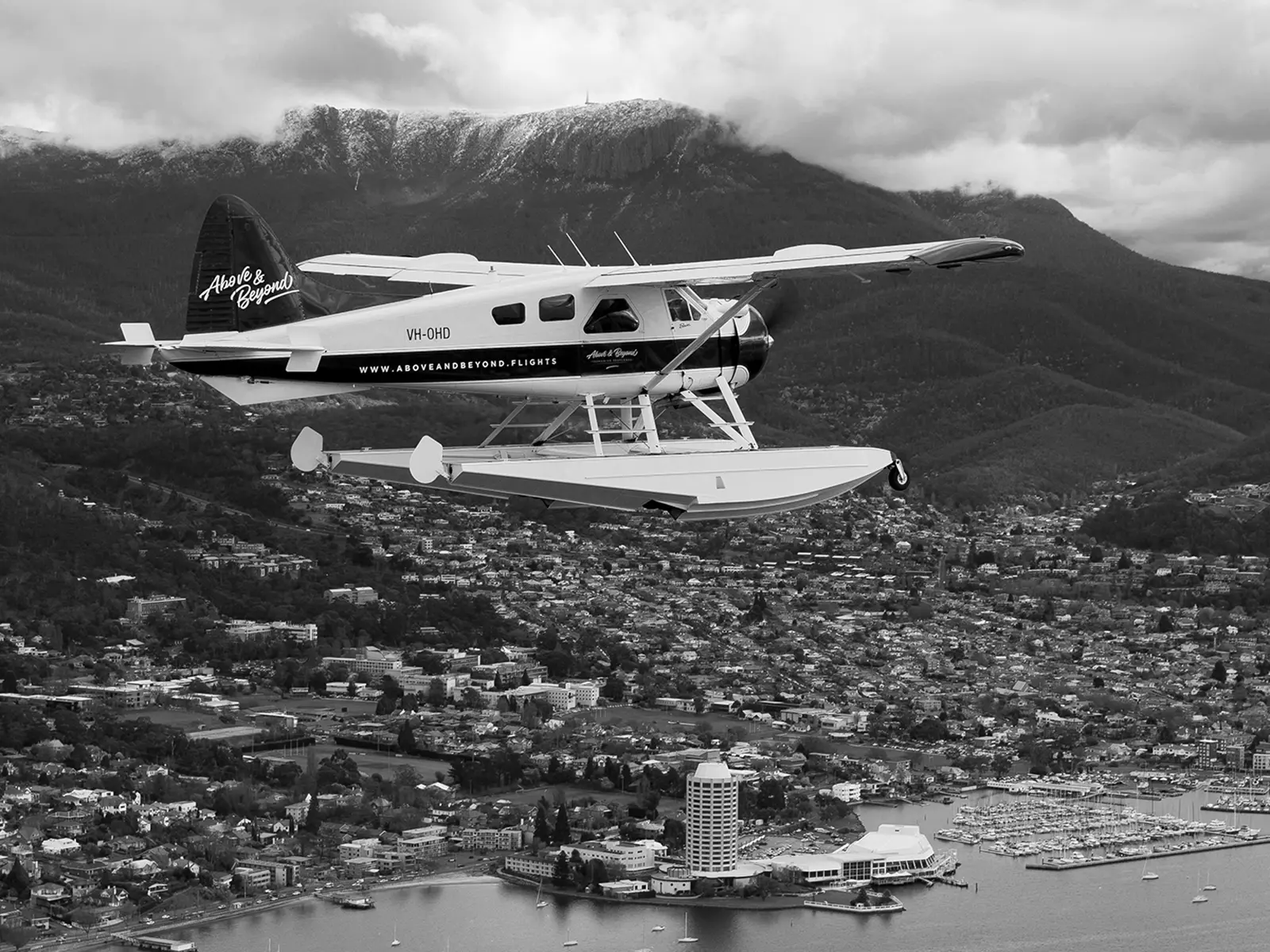 A small seaplane flies in the air above Hobart. Below, the city's waterfront is visible, with snow-dusted mountaintops rising above.