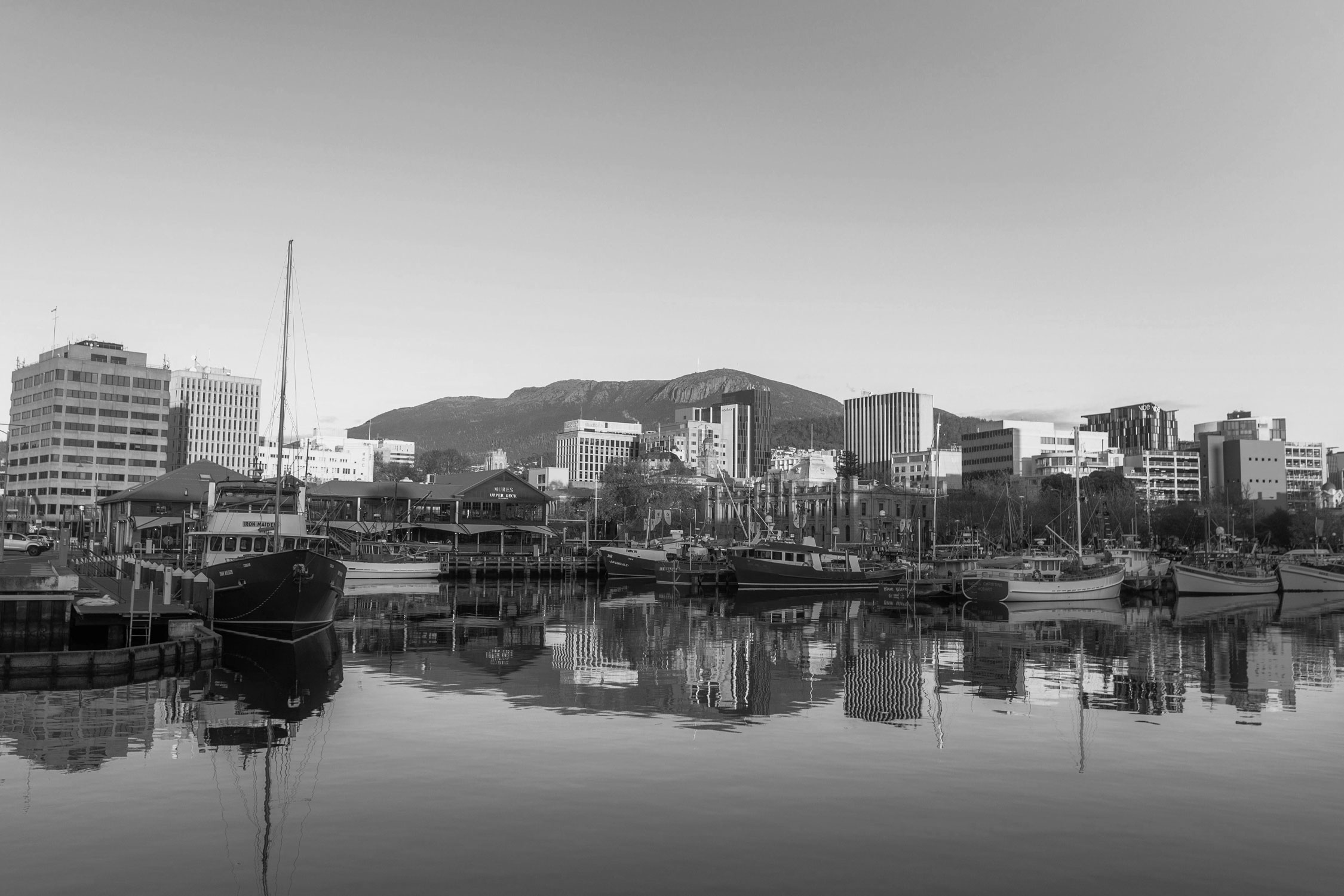 Black and white photo of a harbor scene with boats docked, buildings in the background, and a mountain range behind the city, with calm water reflecting the scene.