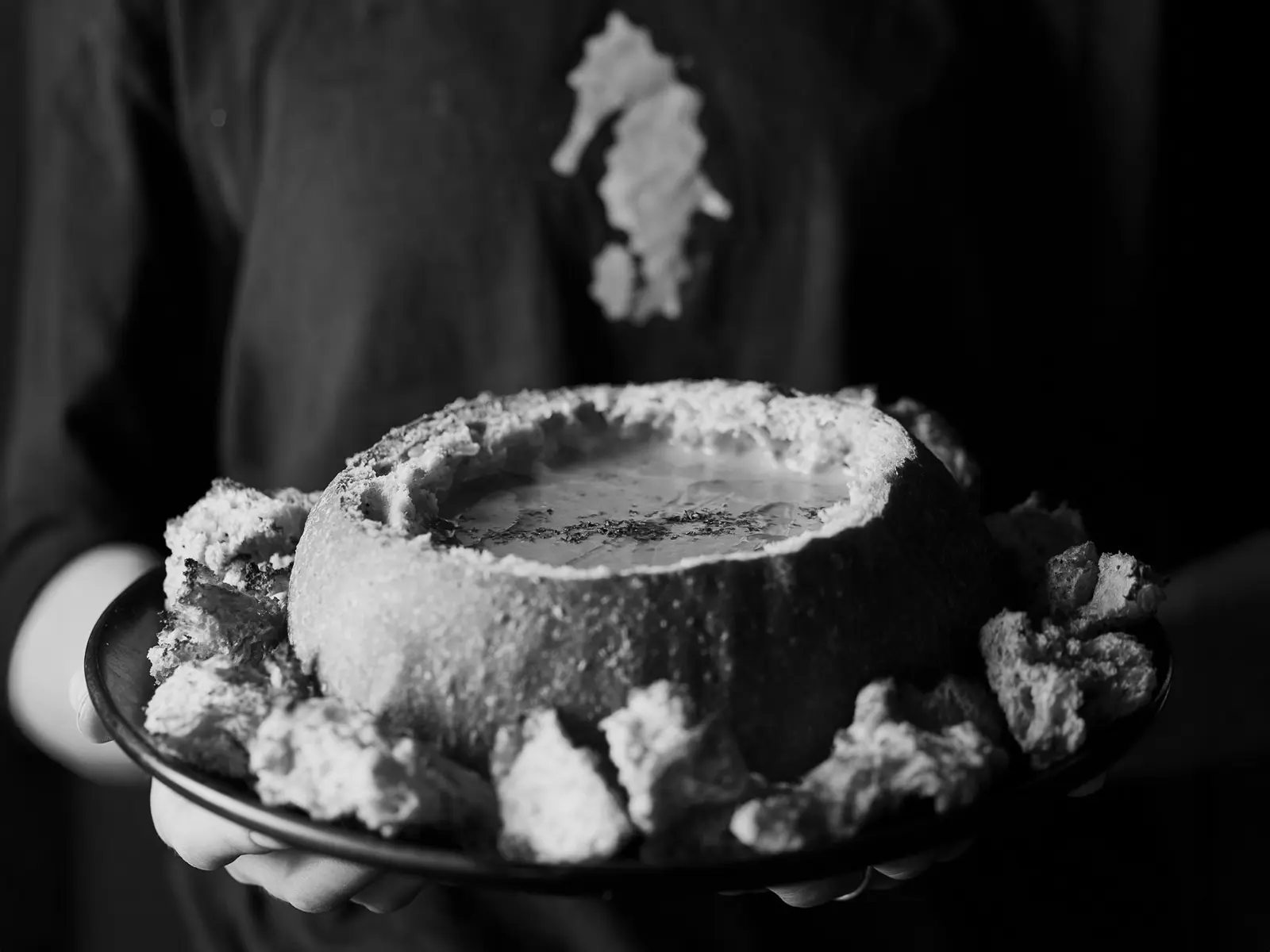 Black and white image of a person holding out a plate holding a cob loaf hollowed out and used as a bowl, filled with soup.