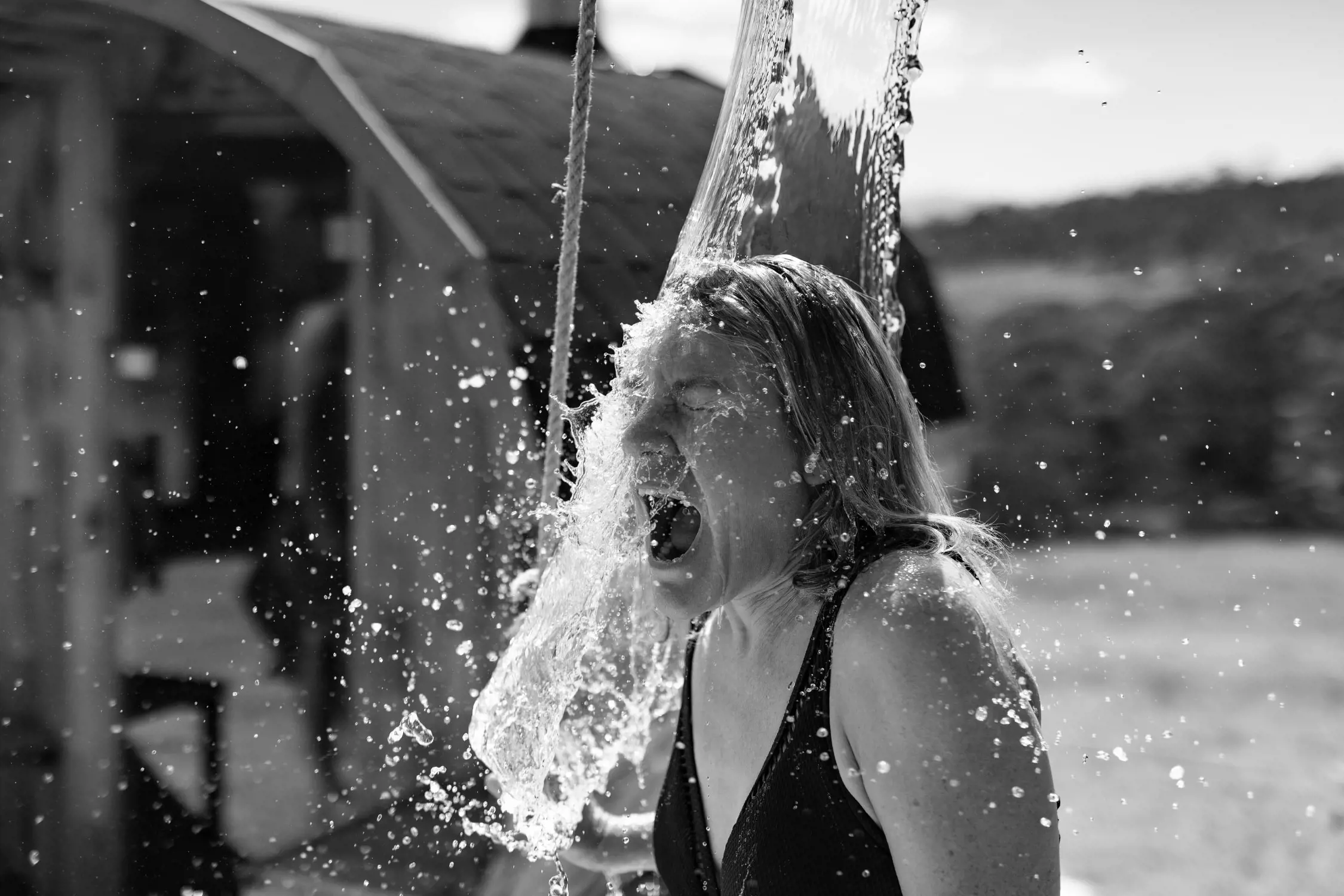 Black and white photo of a woman in a swimsuit being splashed with water on her face, her mouth open wide in reaction.