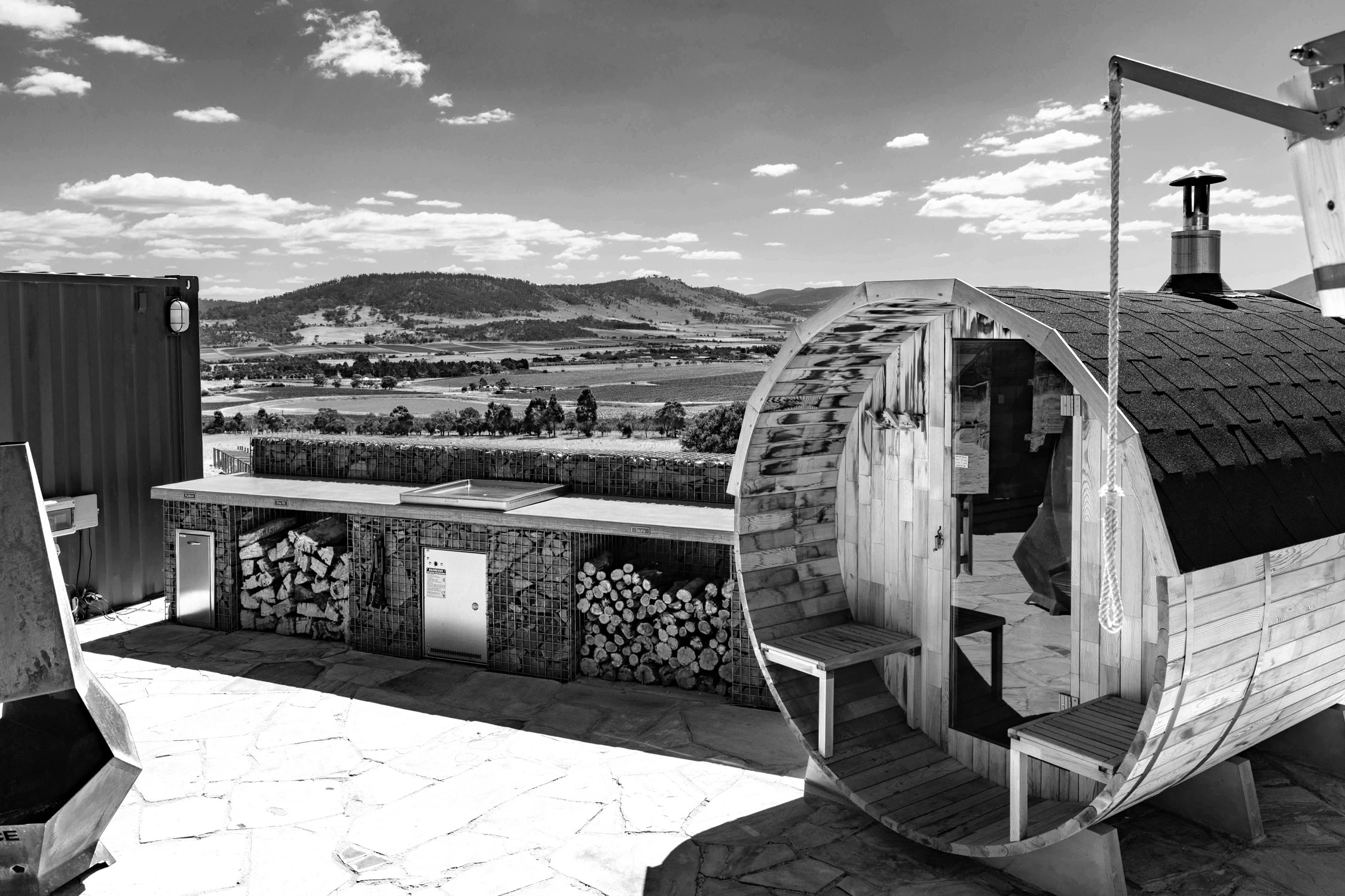 Black and white photo of an outdoor scene showing a wooden barrel-shaped sauna with firewood stacked beside it and a scenic mountain view in the background.