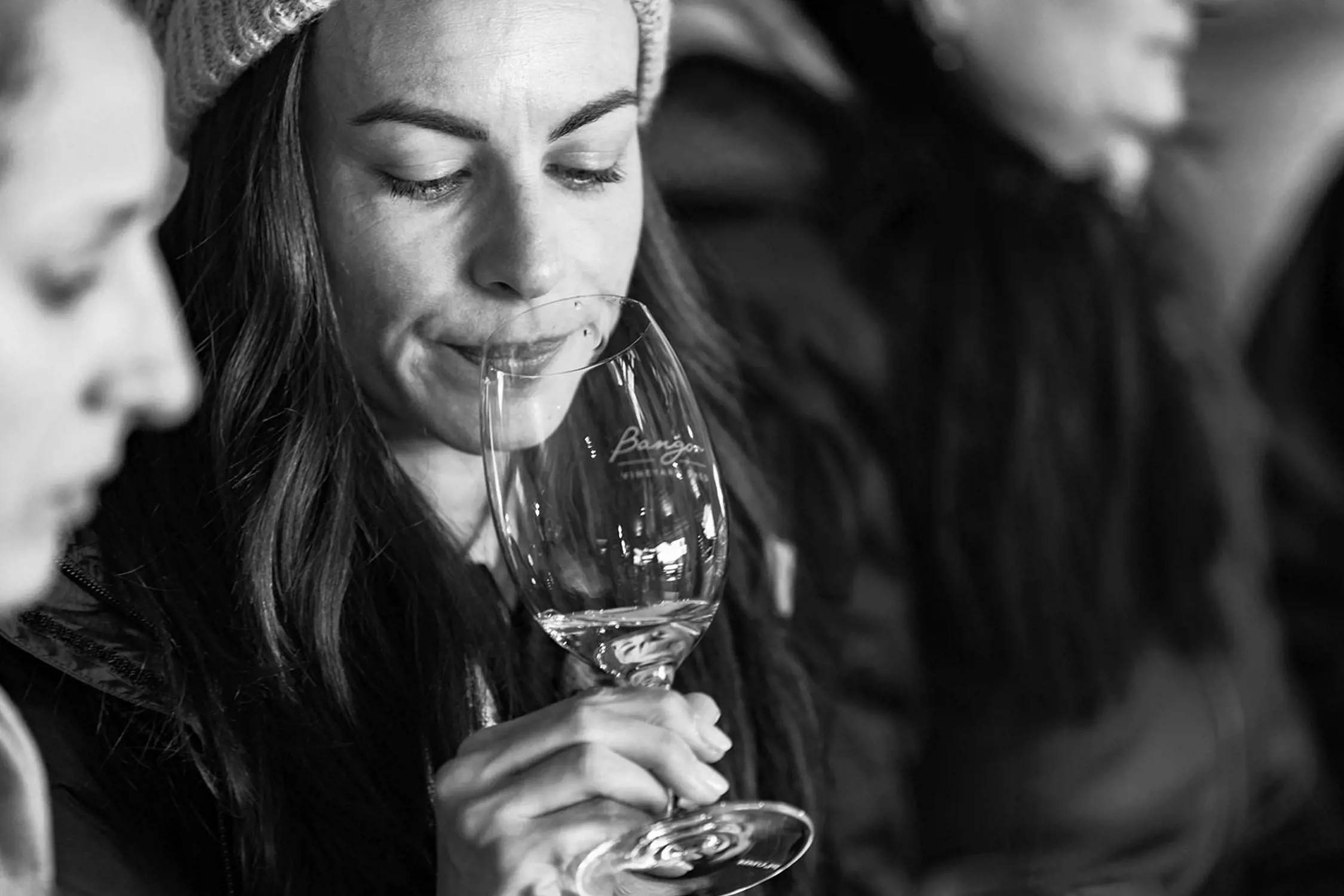 Black and white close-up photo of a woman holding a wine glass near her nose, seemingly smelling or tasting the wine, with other people blurred in the background.