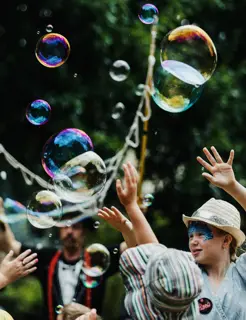 A group of children wearing face paint dance through bubbles at a festival in a park.