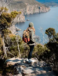 A woman wering a jacket and beanie stands in a clearing on a rocky pathway and looks over ocean and sea cliffs on a sunny day.