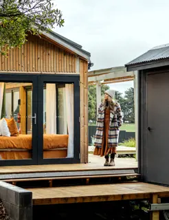 A woman wearing a coat and beanie stands near the entrance to a contemporary wood cabin with large glass doors.