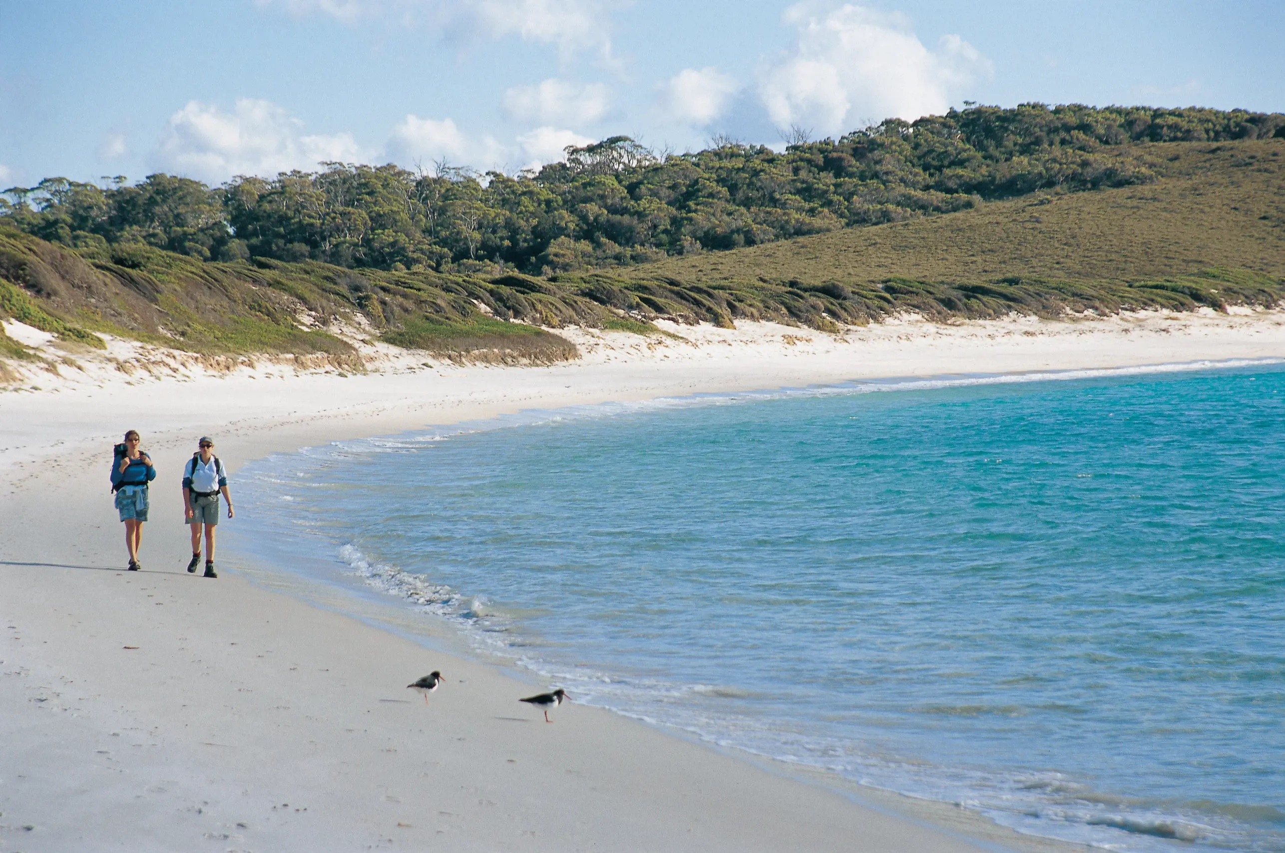 Two people walk along a beach wearing hiking gear and backpacks.