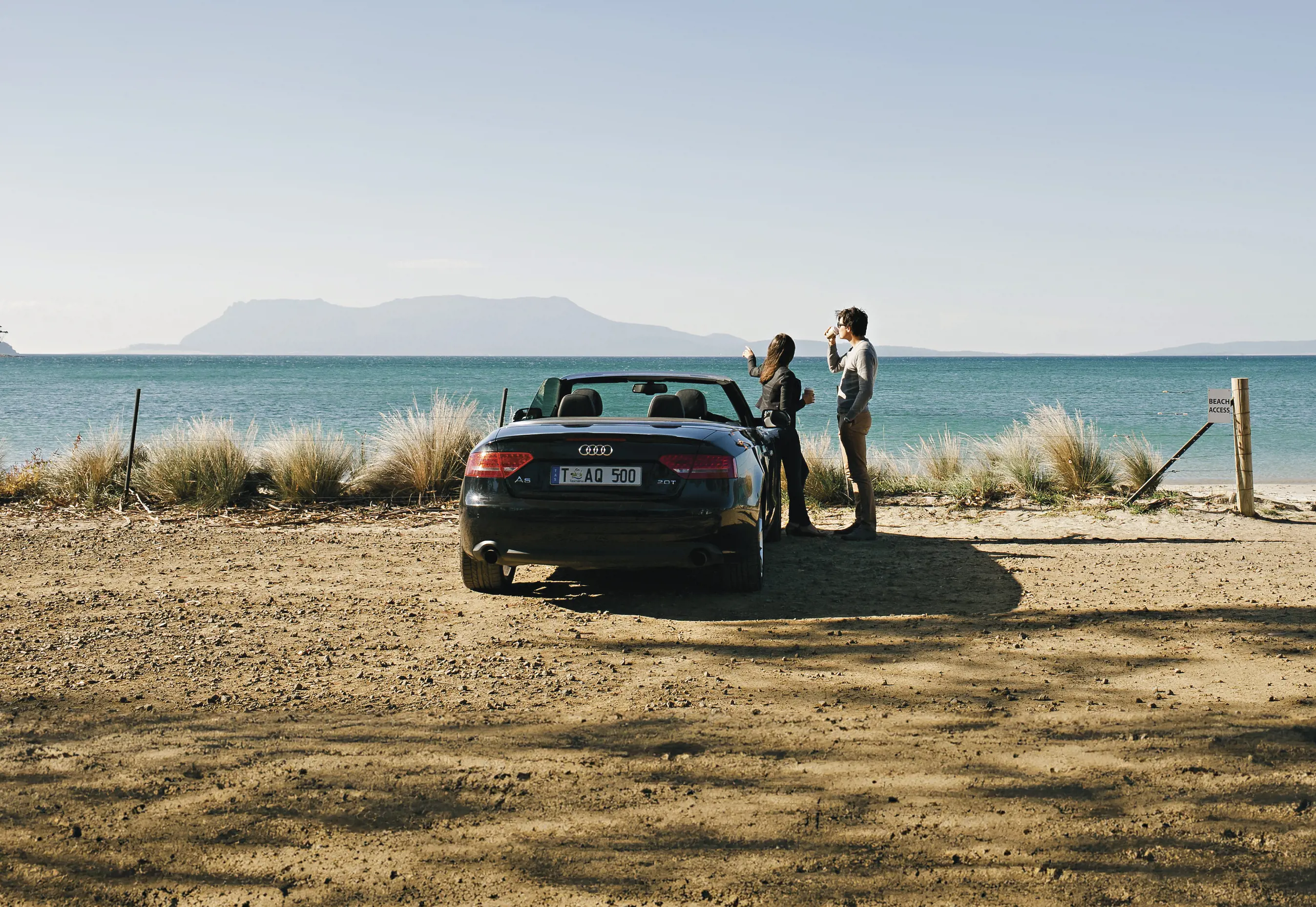 Two people standing next to a convertible car at Spring Beach, Orford. You can see Maria Island in distance.