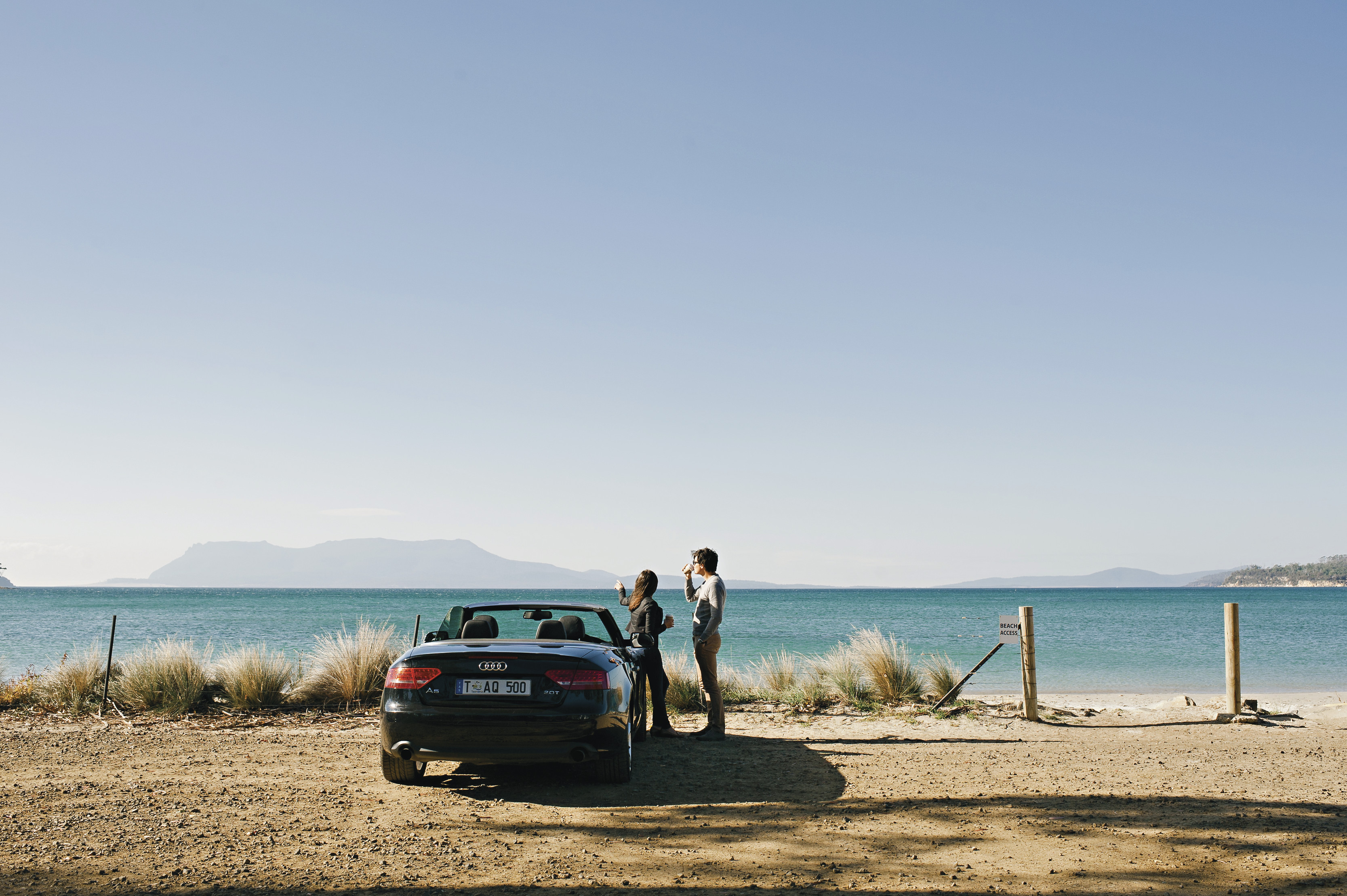 Two people standing next to a convertible car at Spring Beach, Orford. You can see Maria Island in distance. 