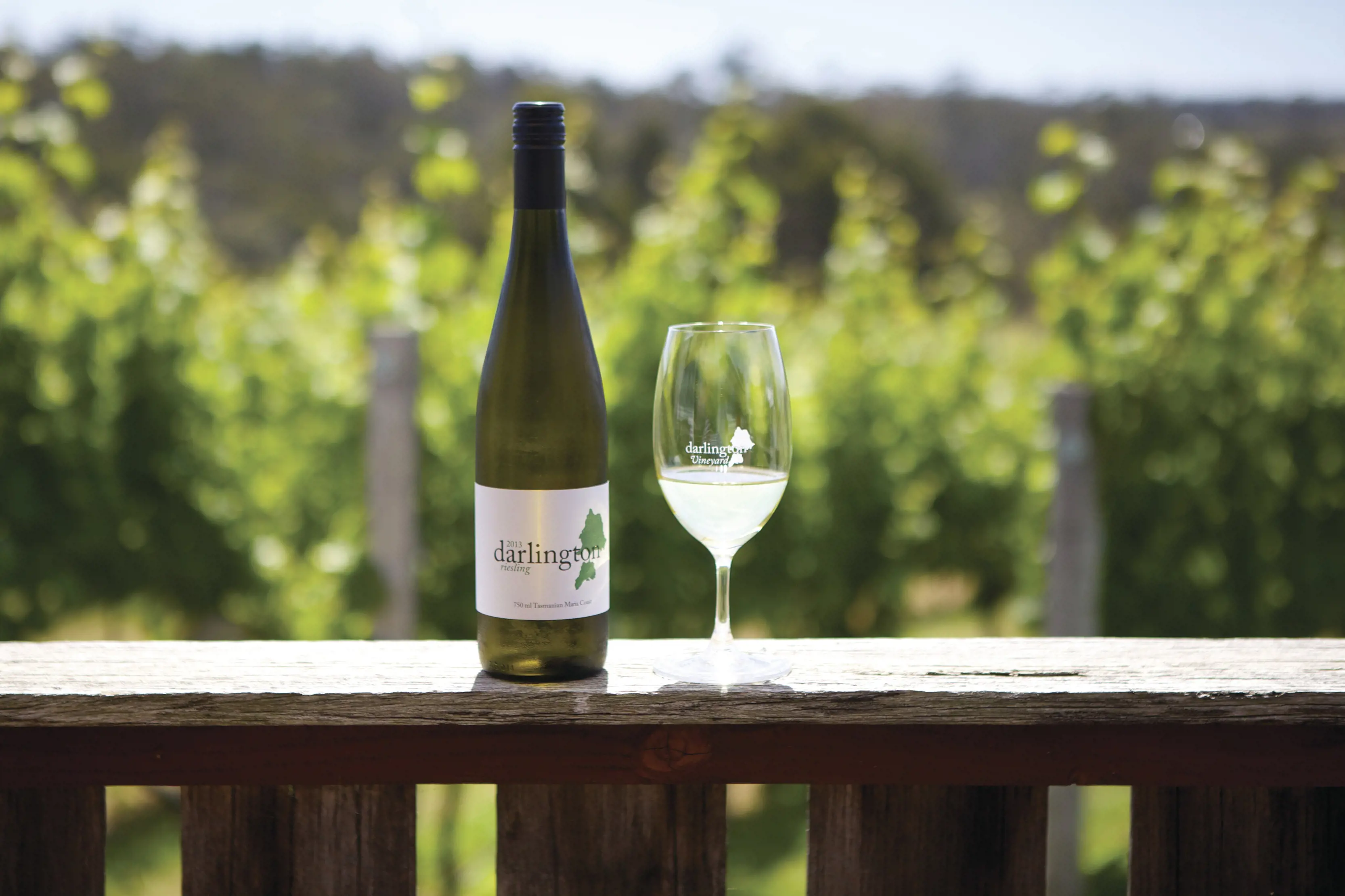 A bottle of Darlington wine and wine glass standing on a wooden frame with greenery in the background. 