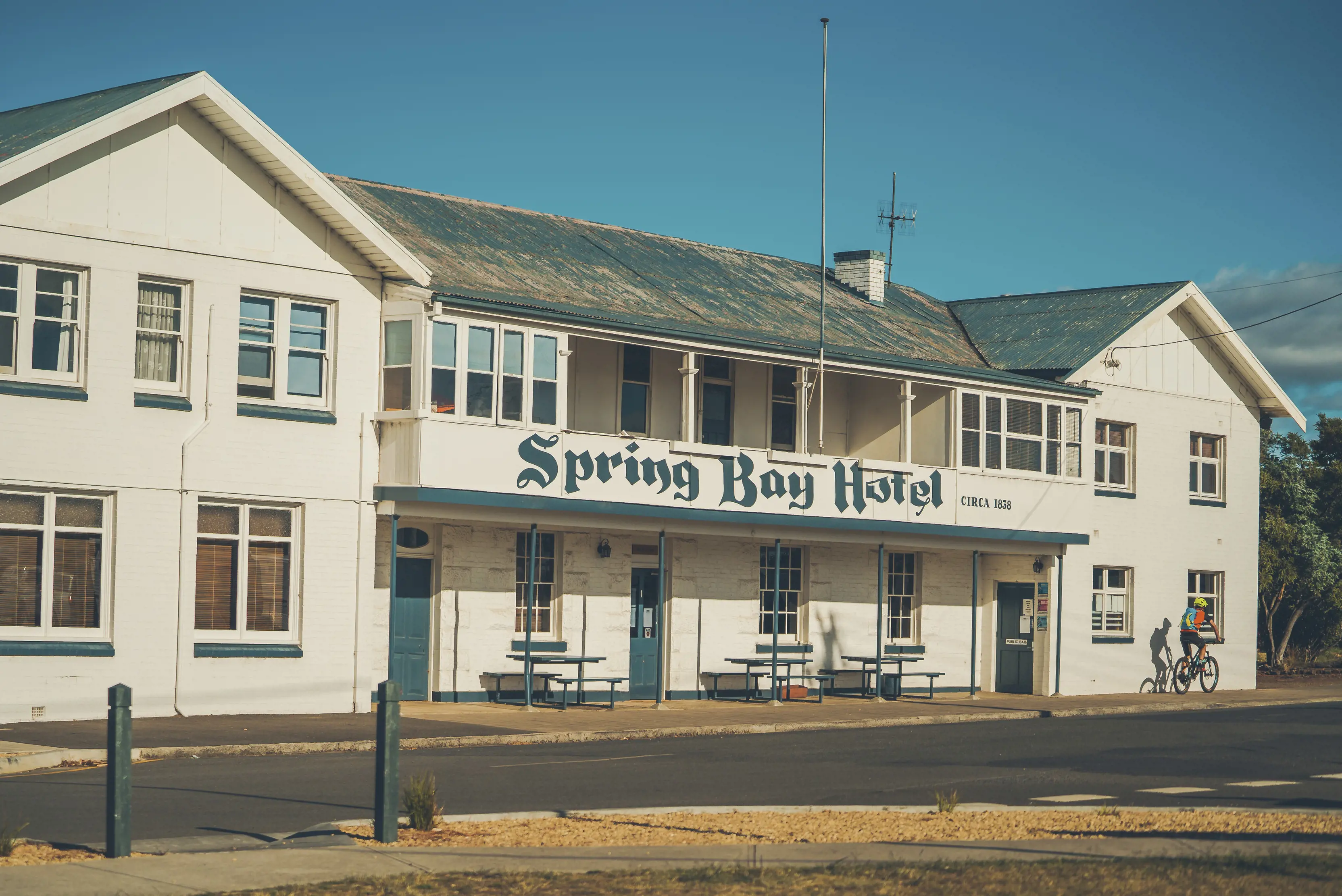 Exterior of Spring Bay Hotel, with signage painted on the hotel front. A cyclist rides past.