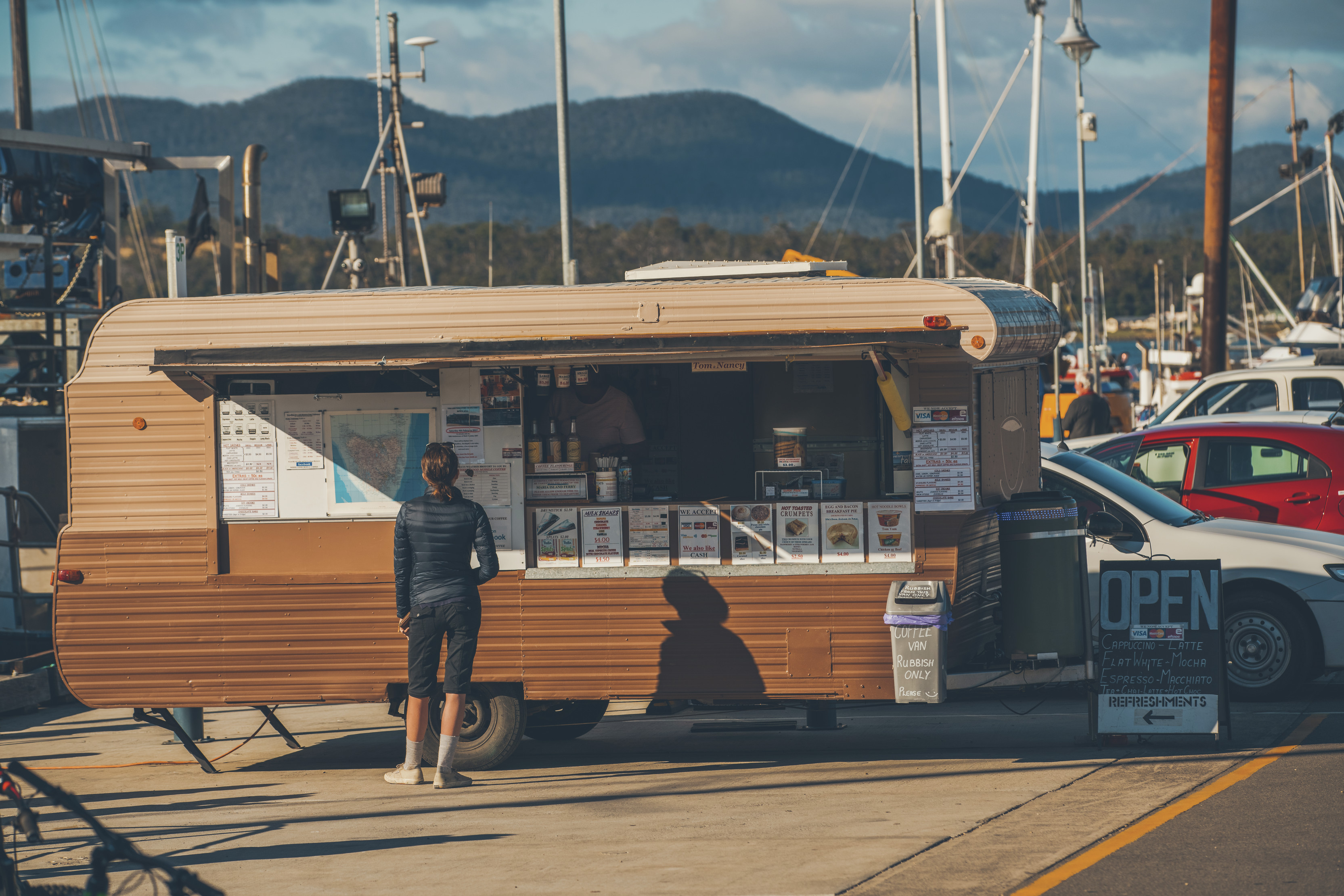 A woman purchases a coffee from the Coffee van, Triabunna. Boats are in the background.
