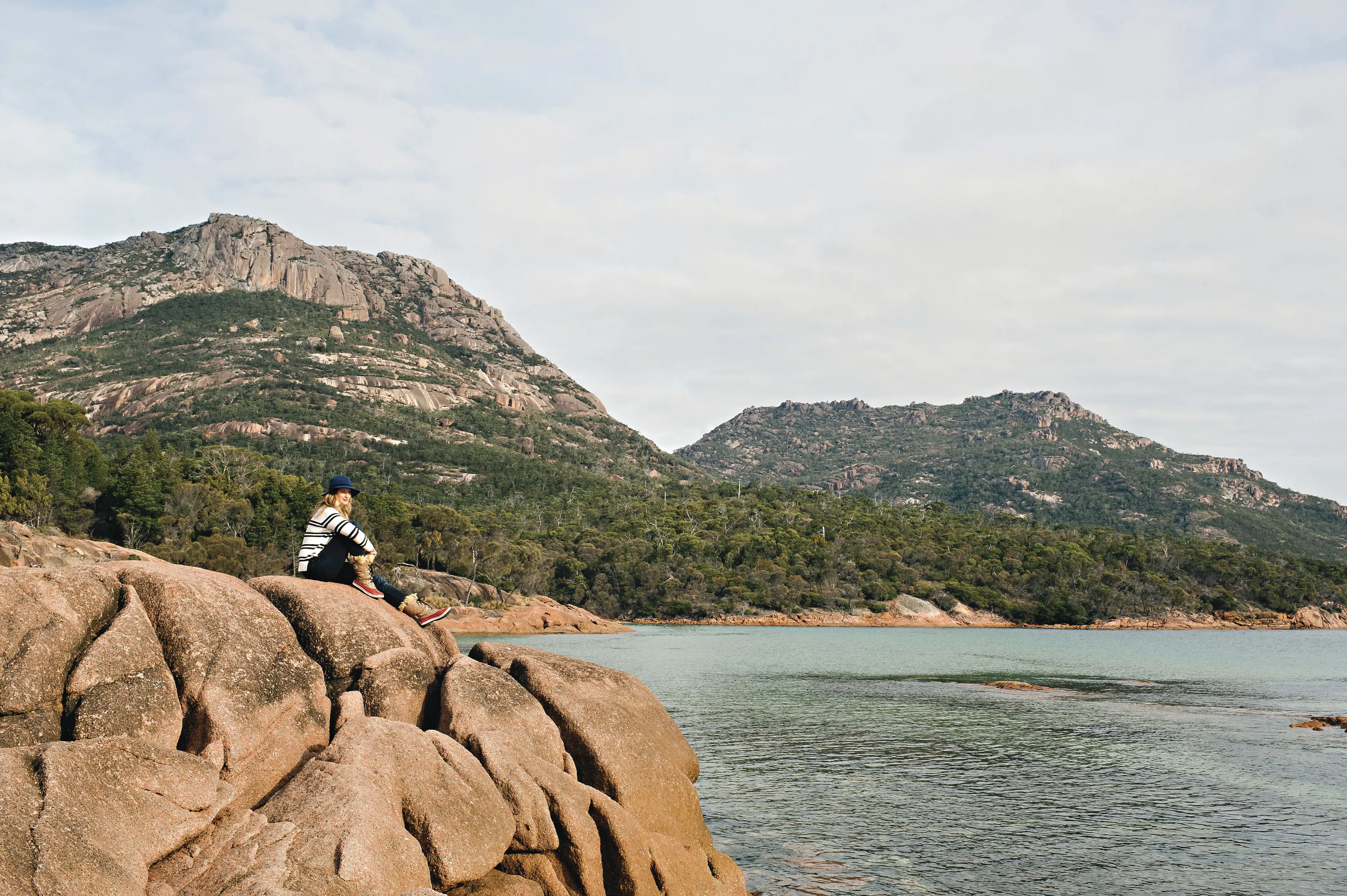 Woman sits on giant rock at Honeymoon Bay, looking out into the ocean. Mountains behind the water. 