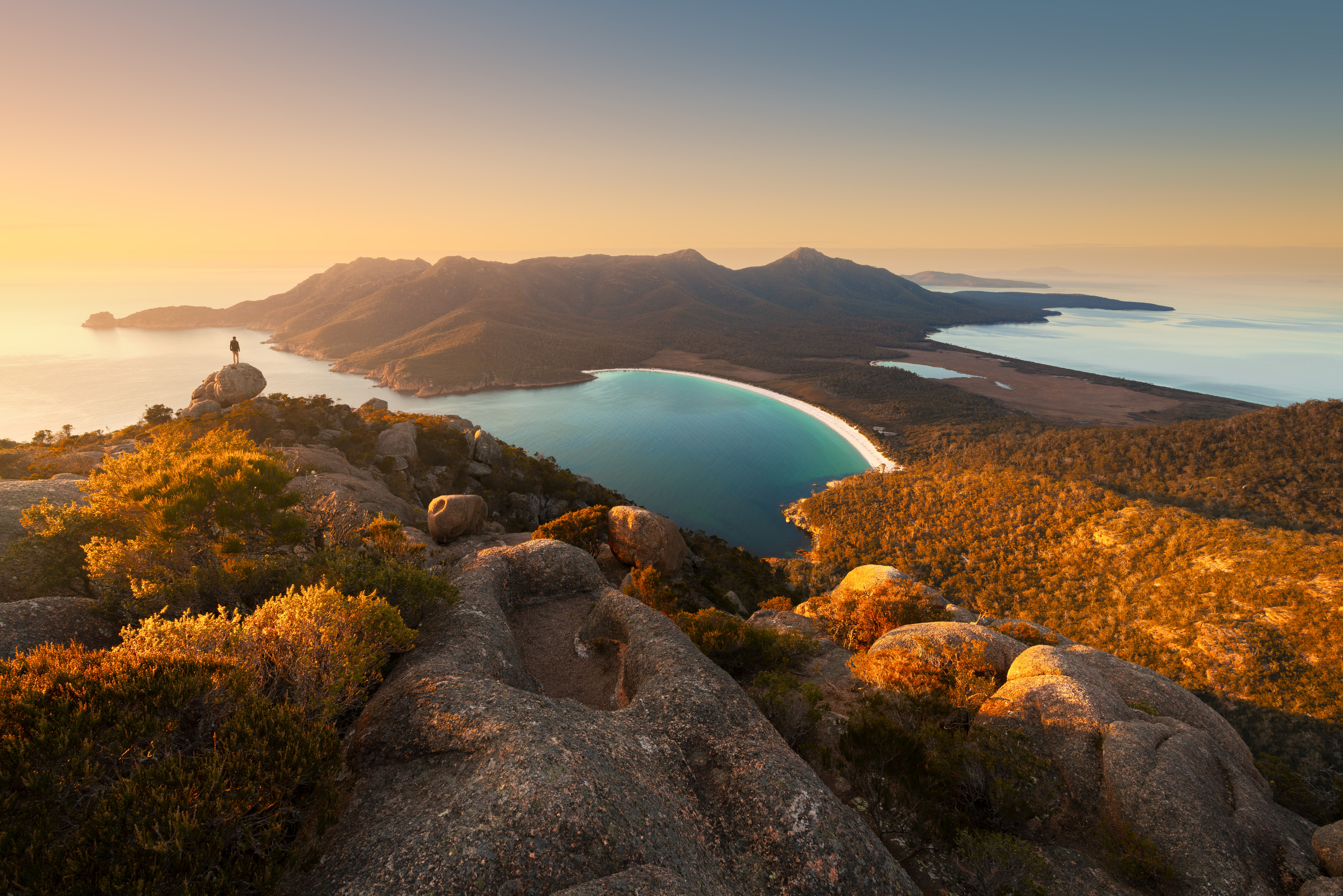 Ariel of Wineglass Bay, a traveller stands on a rock looking at the view. 