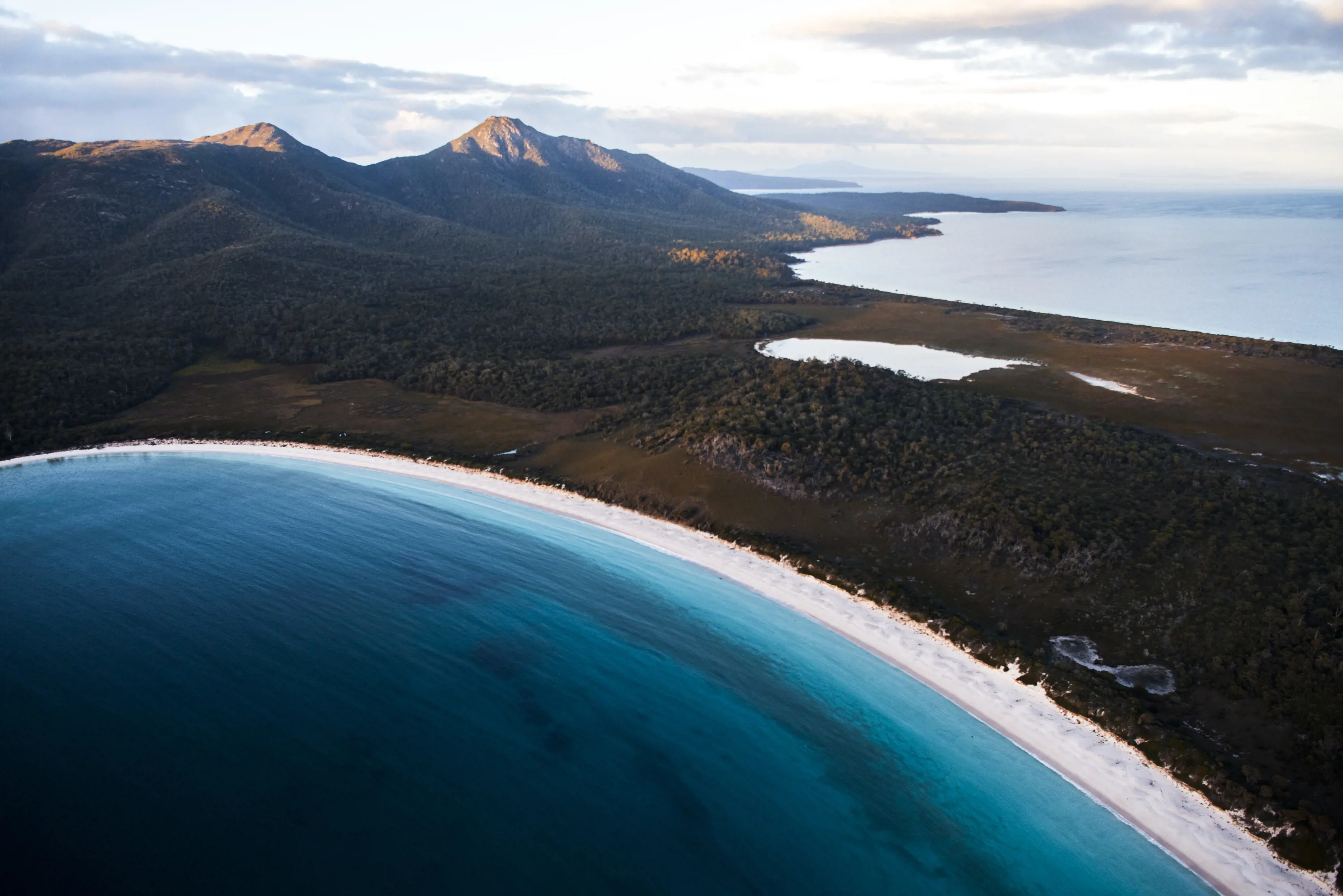 Aerial view of Wineglass Bay beach. Dark blue waters. Mountains in the background and another lake. 