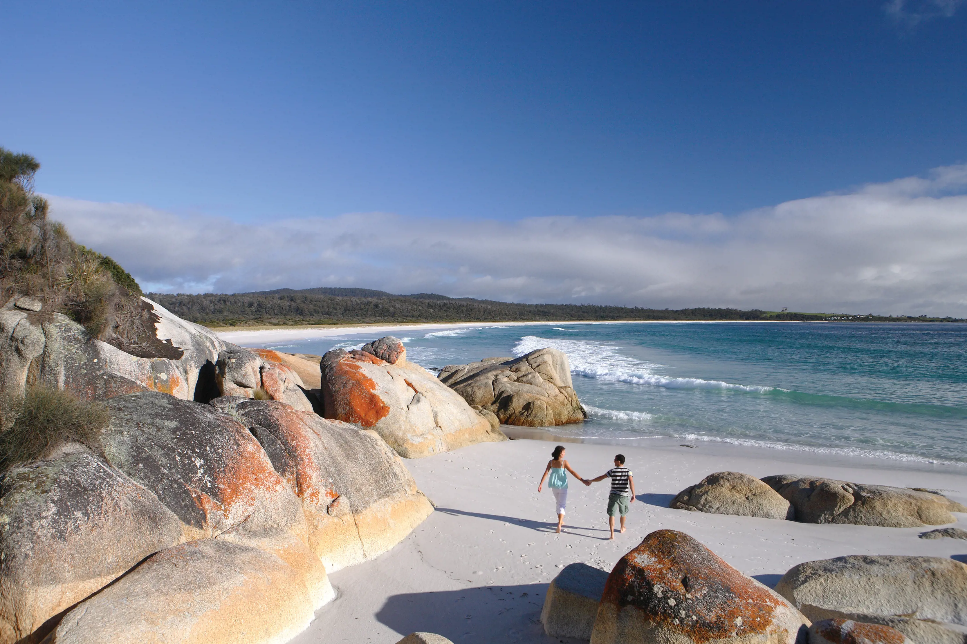 Image of couple walking along the white-sand of Seaton Cove. Surrounded by rocks and ocean.