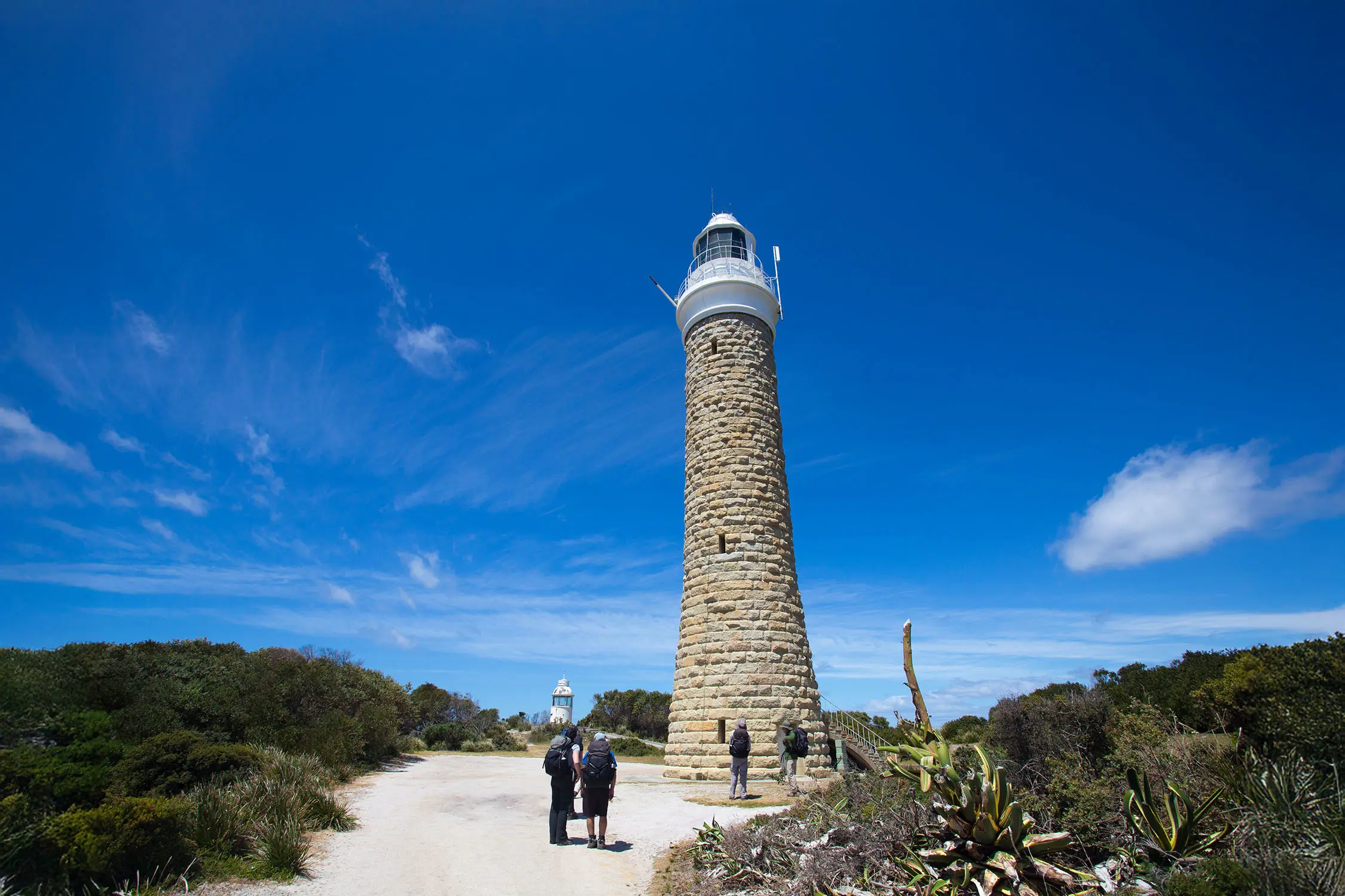 People look up at the tall sandstone lighthouse, surrounded by bush.