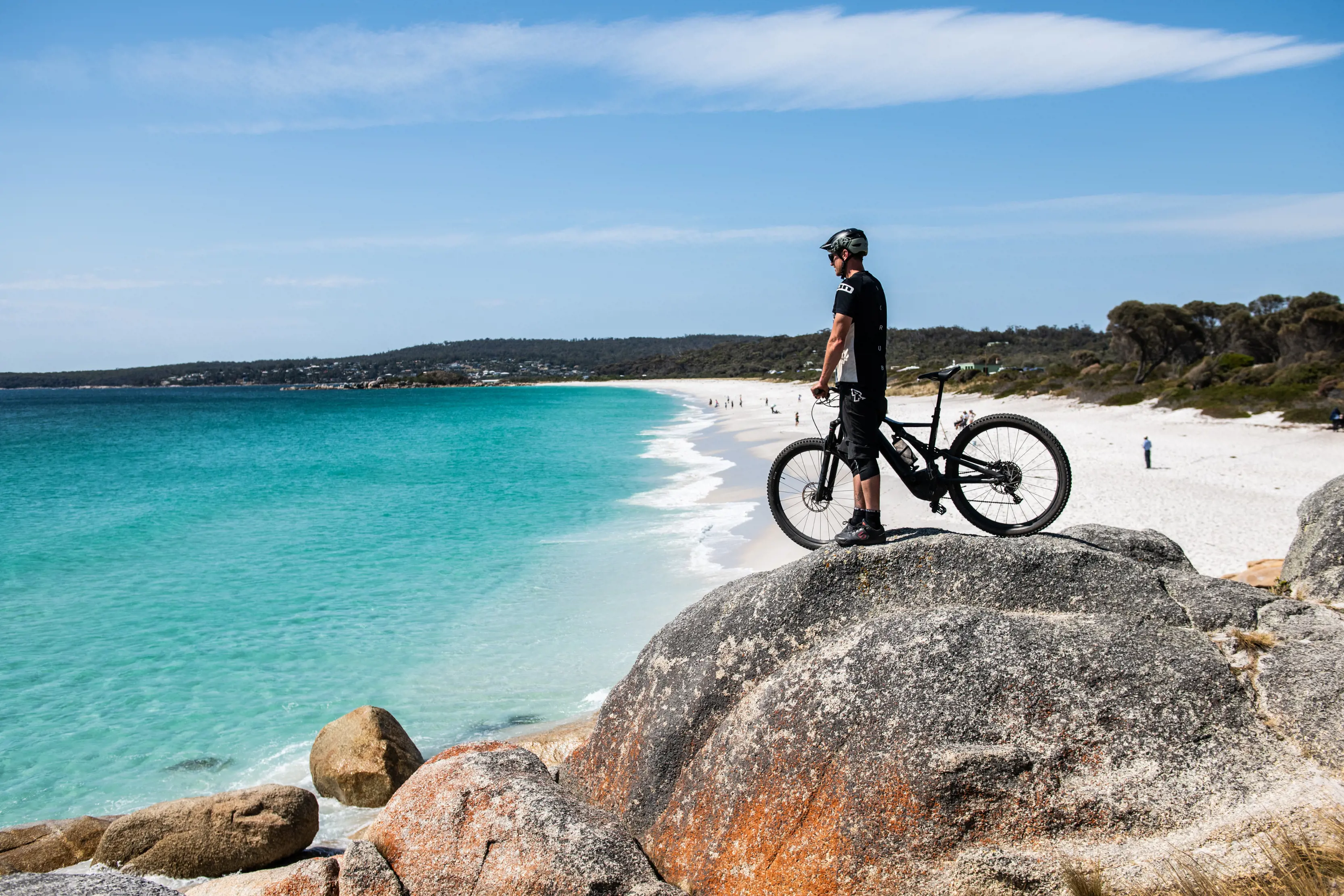 A mountain bike rider stands on the orange coloured boulders of the Bay Of Fires, looking out to sea.