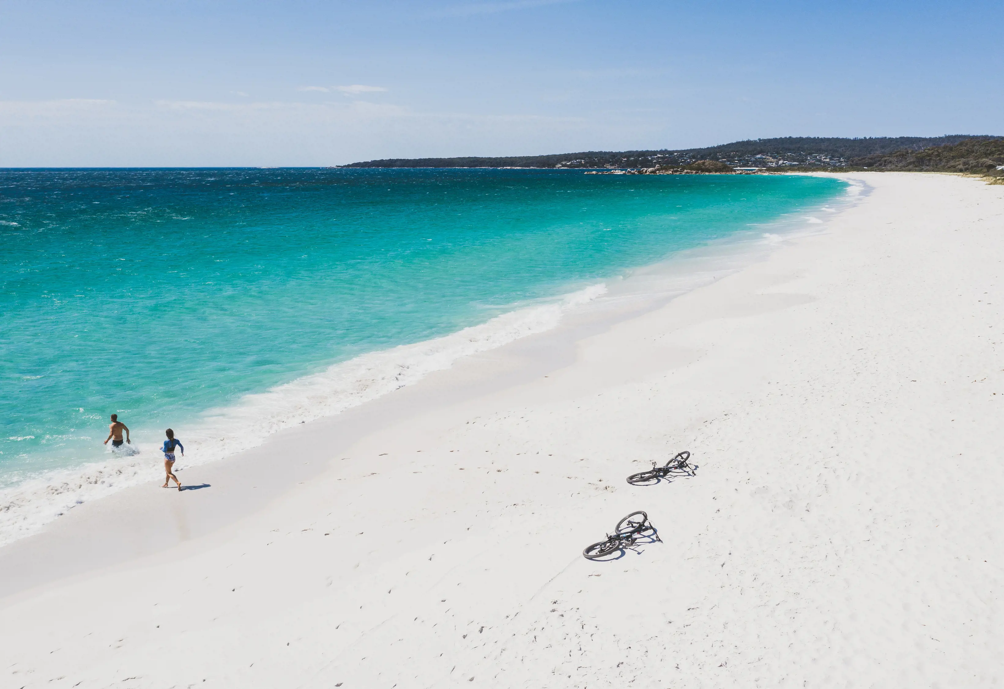 An aerial shot shows two bike riders going for a swim, while their bikes rest on the beach. Swimcart Beach, Bay Of Fires.