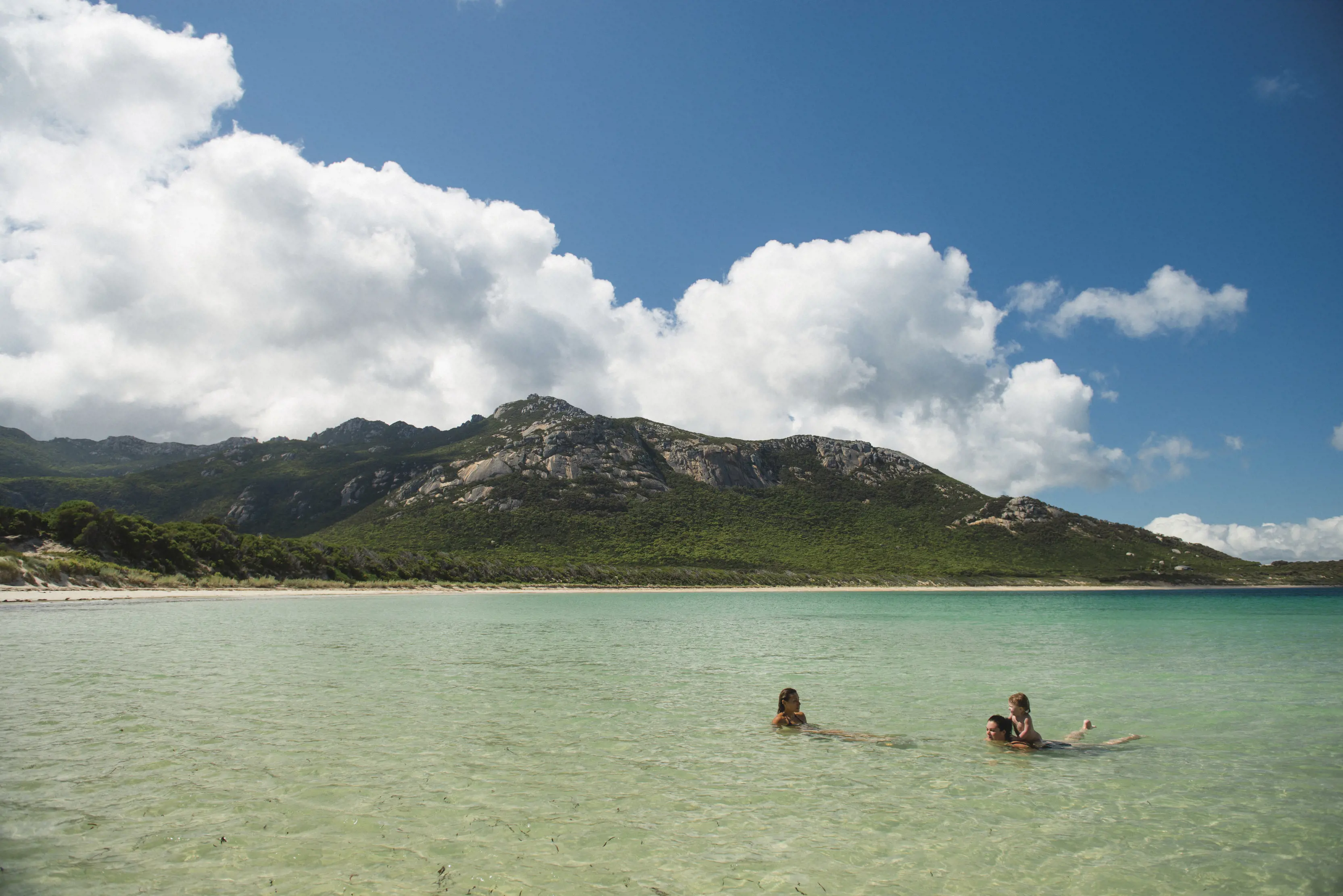 Parents and kid swimming at Trousers Point Beach, with crystal clear waters, wide expanses of white sand, and a mountain rising from the ocean.