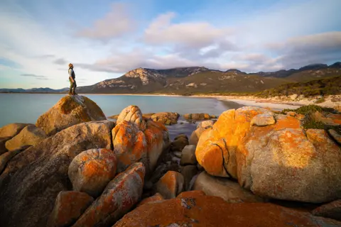 Breathtaking image of Fotheringate Bay, Flinders Island, of a man standing on a large rock, looking over the stunning coastline.