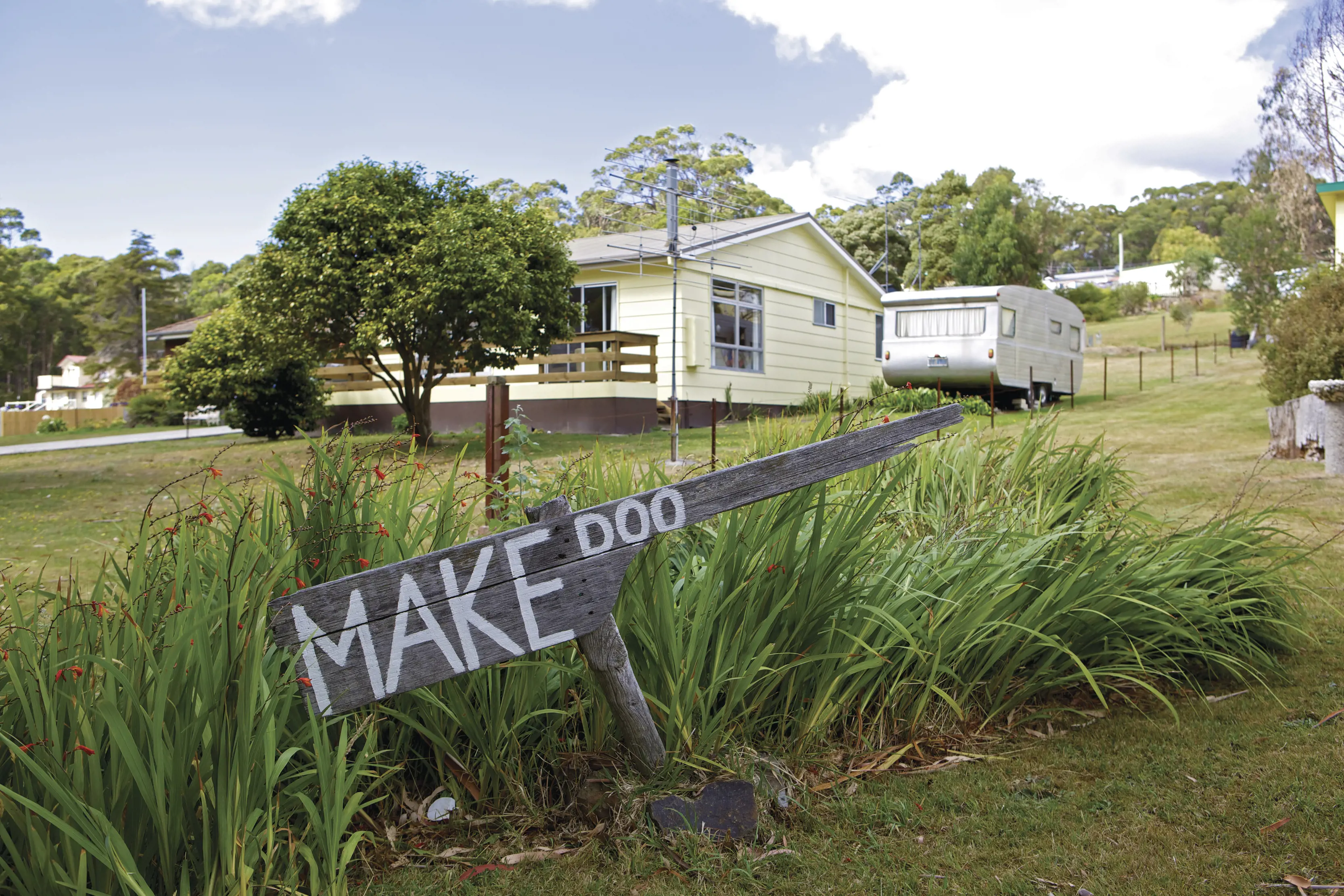 Old wooden sign that says Make Doo, sitting in front of a white house in Make Doo - Doo Town