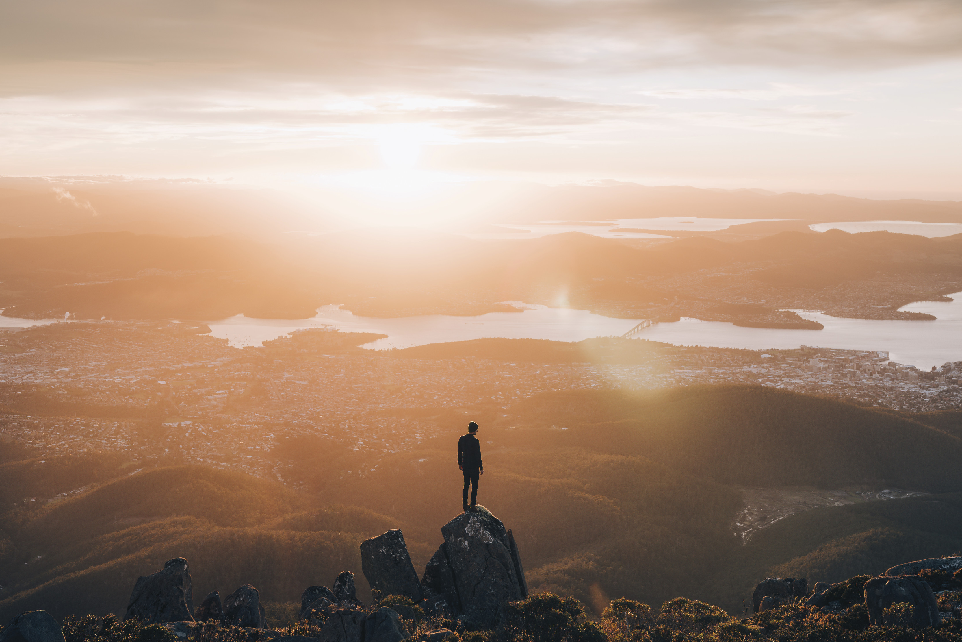 Incredible, dramatic image of a person standing on a rock, on the Summit of kunanyi / Mt Wellington, overlooking the vast landscape during sunrise. 