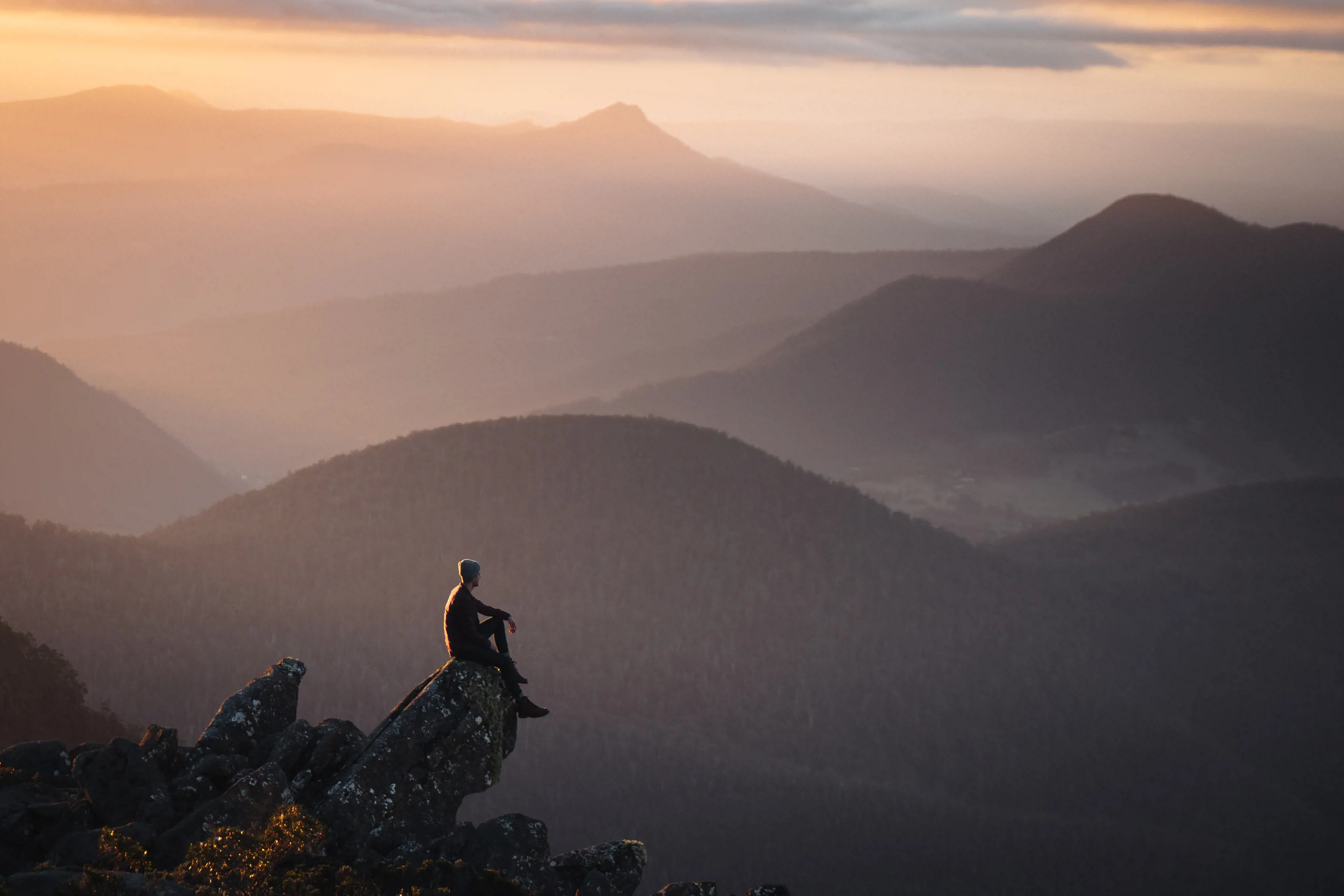 Incredible, dramatic image of a person sitting on a rock, on the Summit of kunanyi / Mt Wellington, overlooking the vast landscape during sunrise.