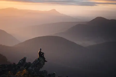 Incredible, dramatic image of a person sitting on a rock, on the Summit of kunanyi / Mt Wellington, overlooking the vast landscape during sunrise.