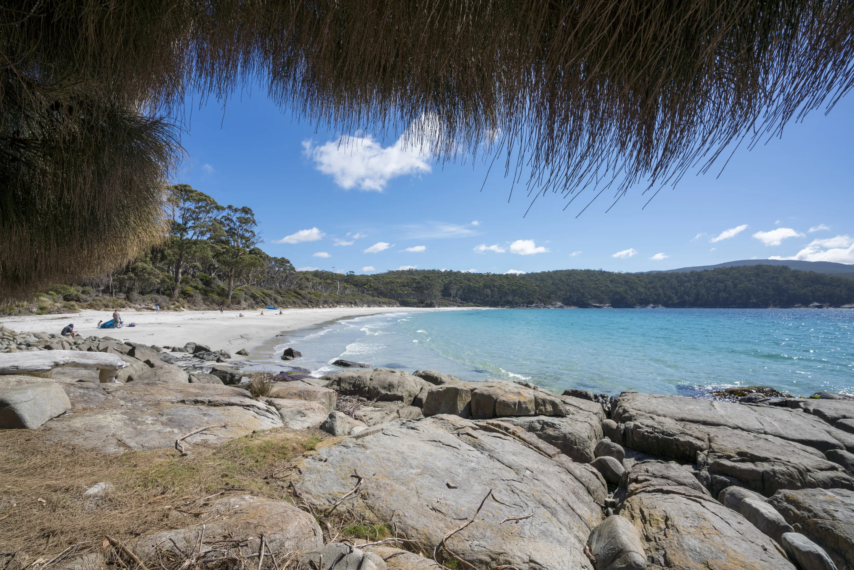 Looking out over rocks, with a sandy beach and bright blue sea in the distance at Fortescue Bay, part of the Three Capes Track.