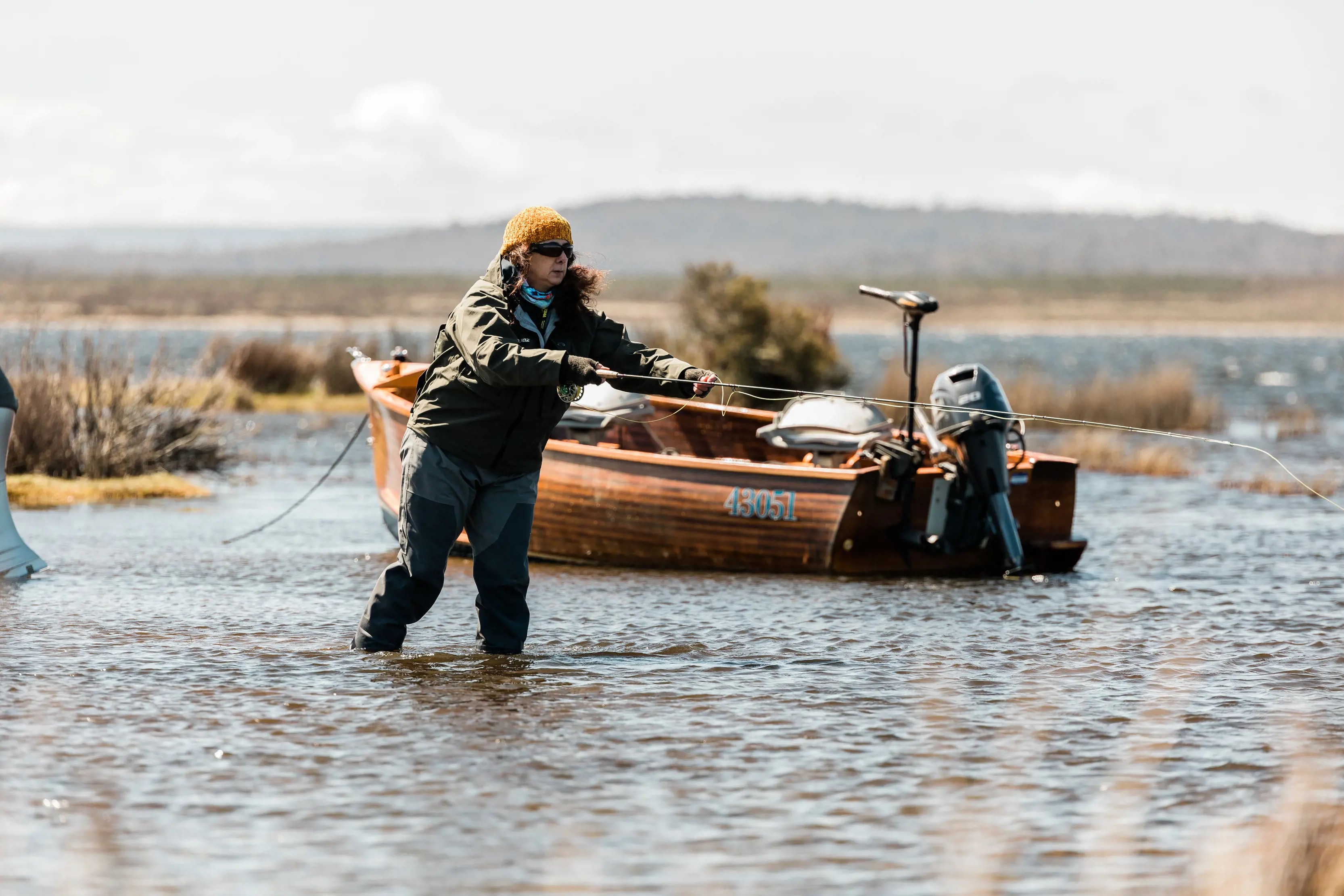 A person stands in the shallow waters fly fishing at Little Pine Lagoon, a boat is parked still behind them.