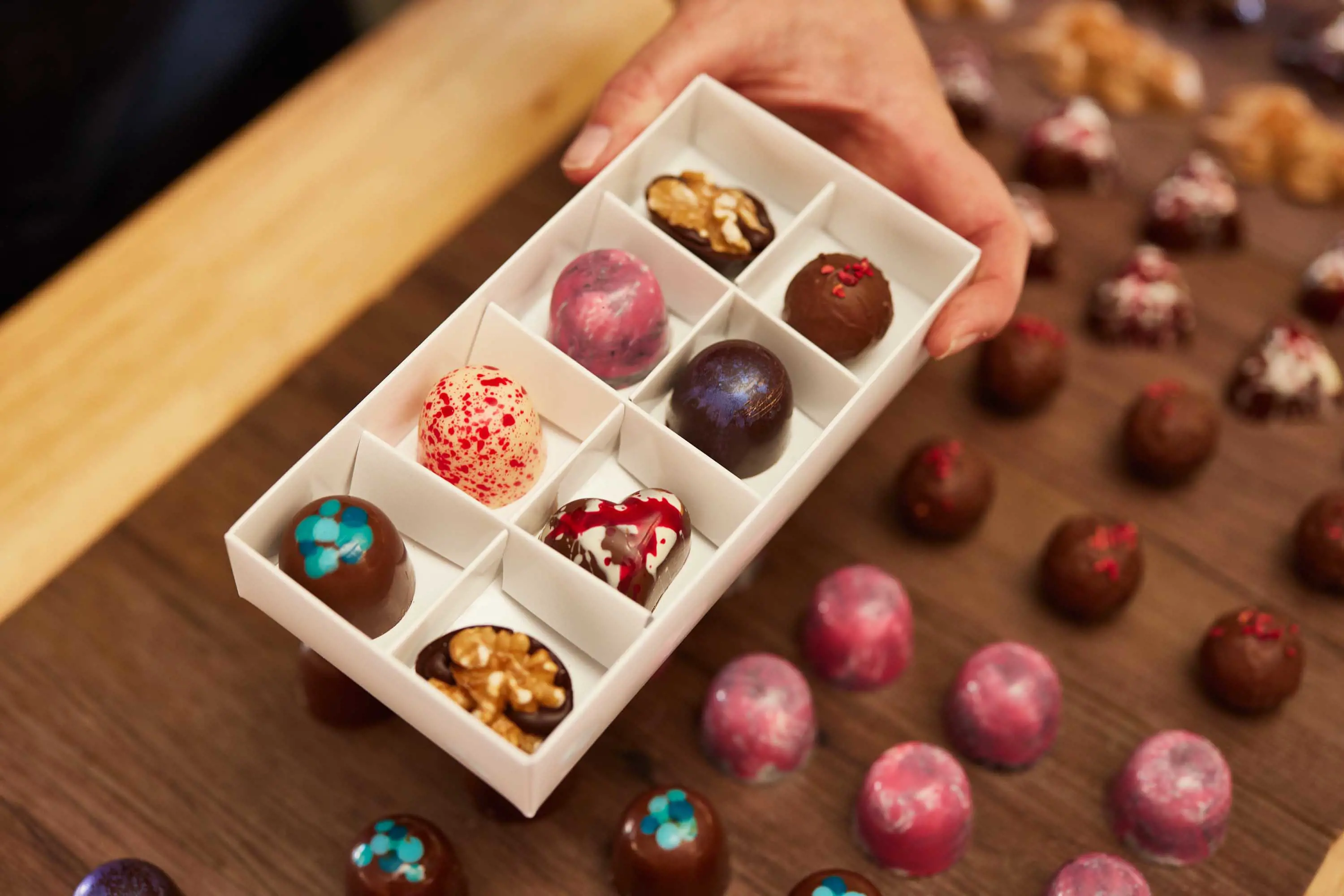 Eight, round,  multi-coloured chocolates are displayed in a white box and held above a cabinet filled with other chocolate varieties.